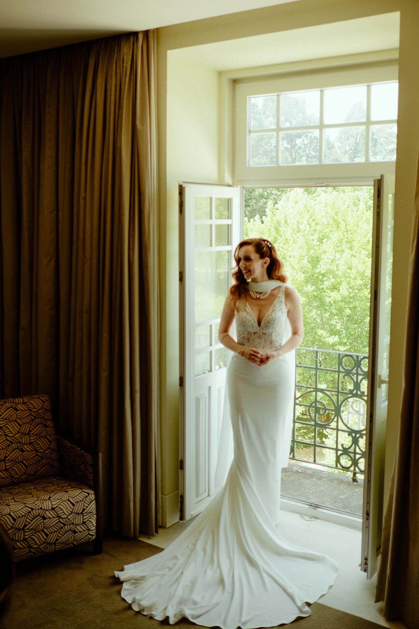 Bride in white lace wedding gown with train stands in open French doors with balcony view.
