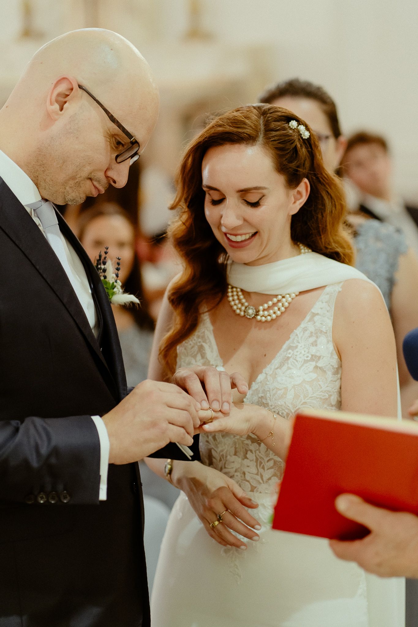 Groom placing wedding ring on bride's finger during ceremony, bride smiling in white lace dress with pearl necklace.