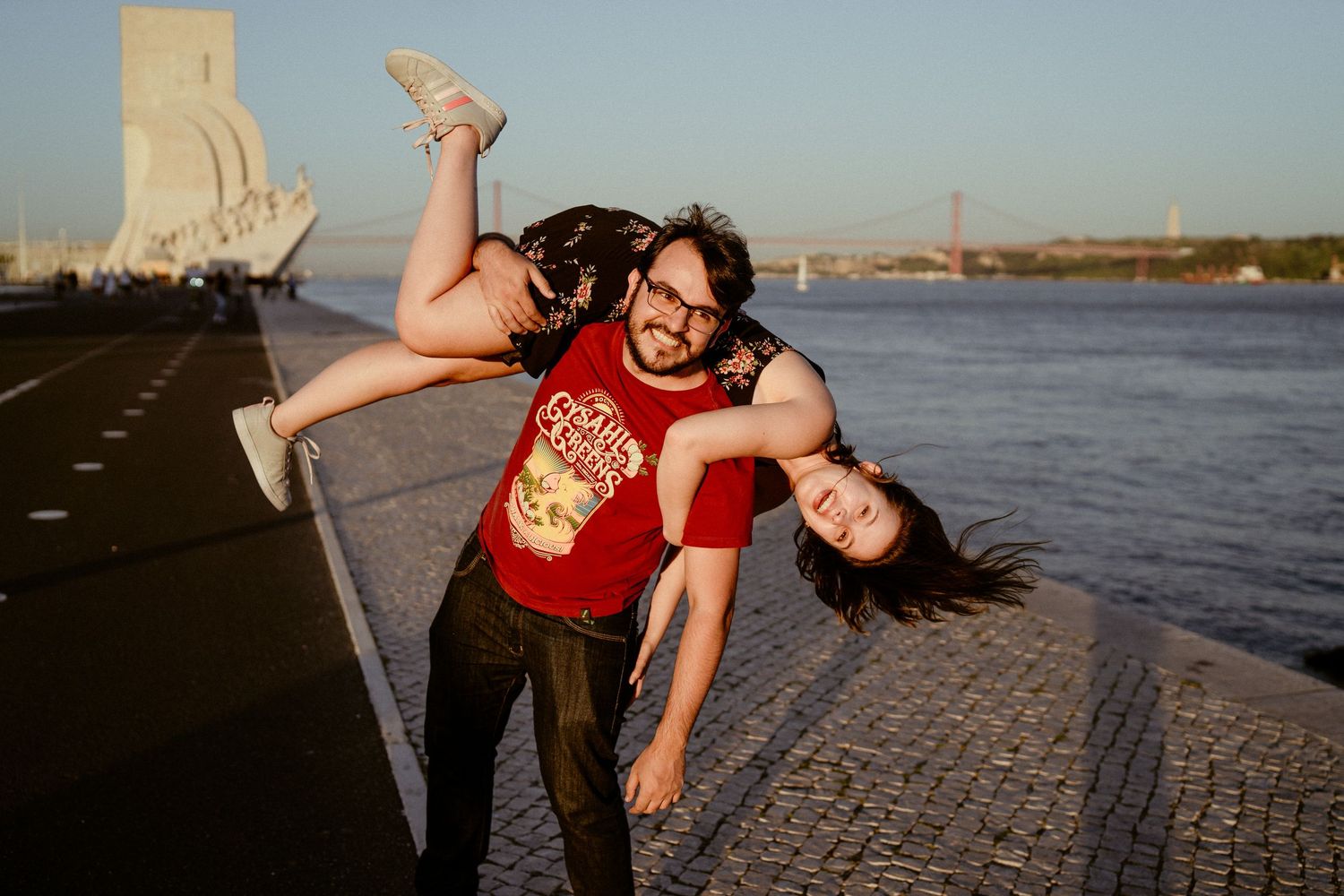 A couple poses playfully near the water during a sunset engagement photo session at a waterfront location.