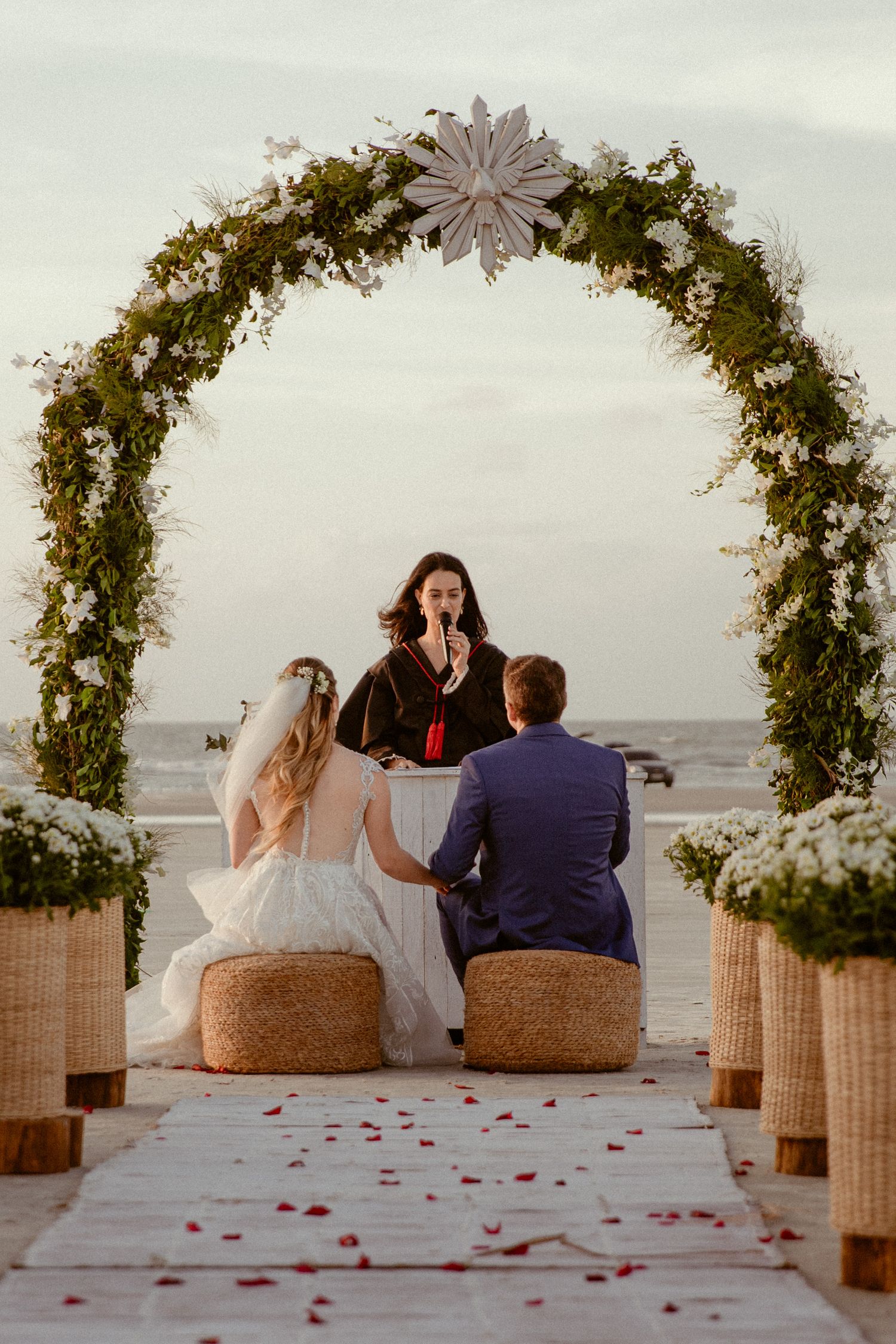 Romantic seaside ceremony setup with floral arches and rose petals scattered on the walkway at sunset.