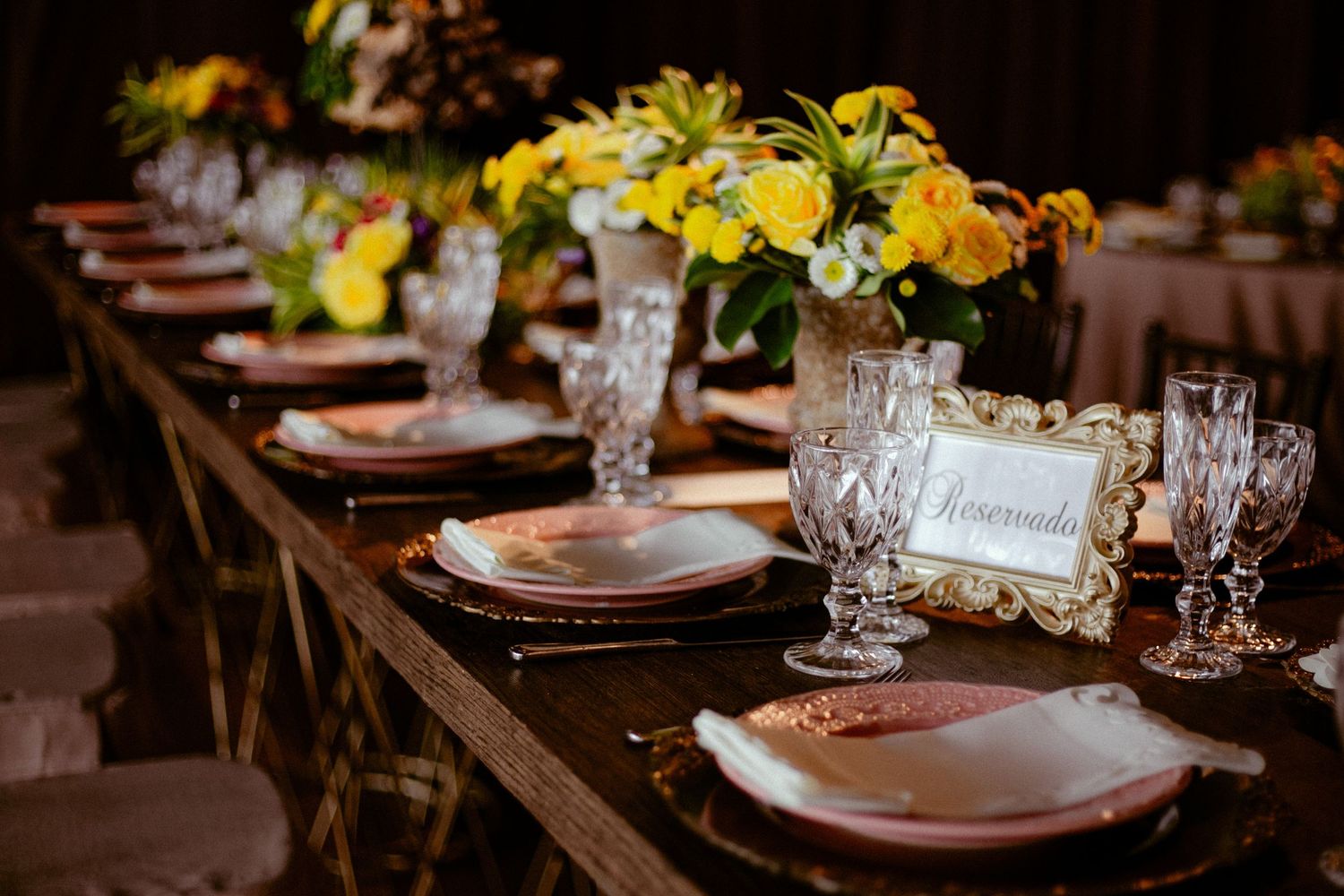 Elegant wedding reception table setup with rustic wood tables, white plates, and yellow floral centerpieces.
