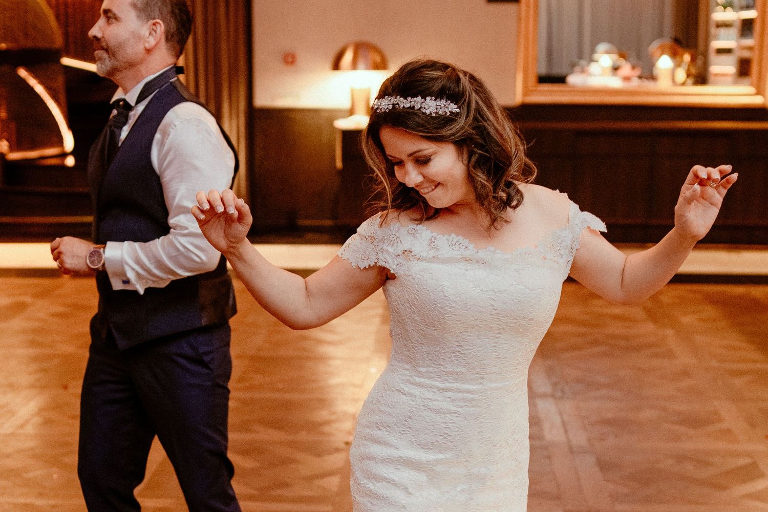 A couple dances together at a wedding reception on a wooden dance floor with warm lighting.