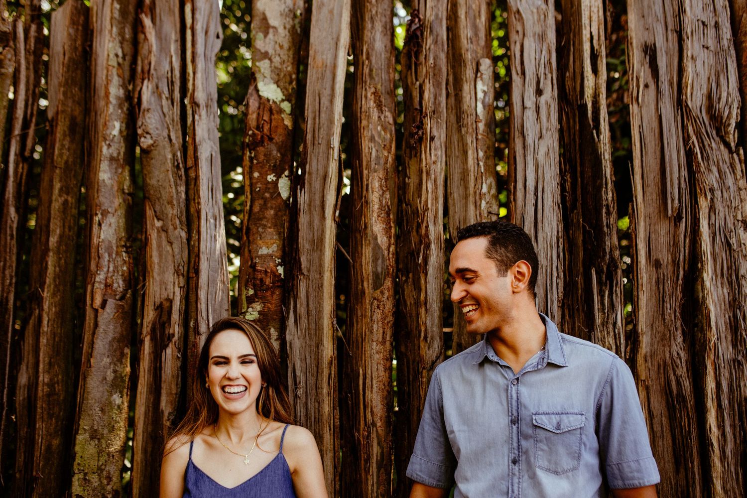 Couple laughing together against a rustic wooden fence background during a warm outdoor photo session.
