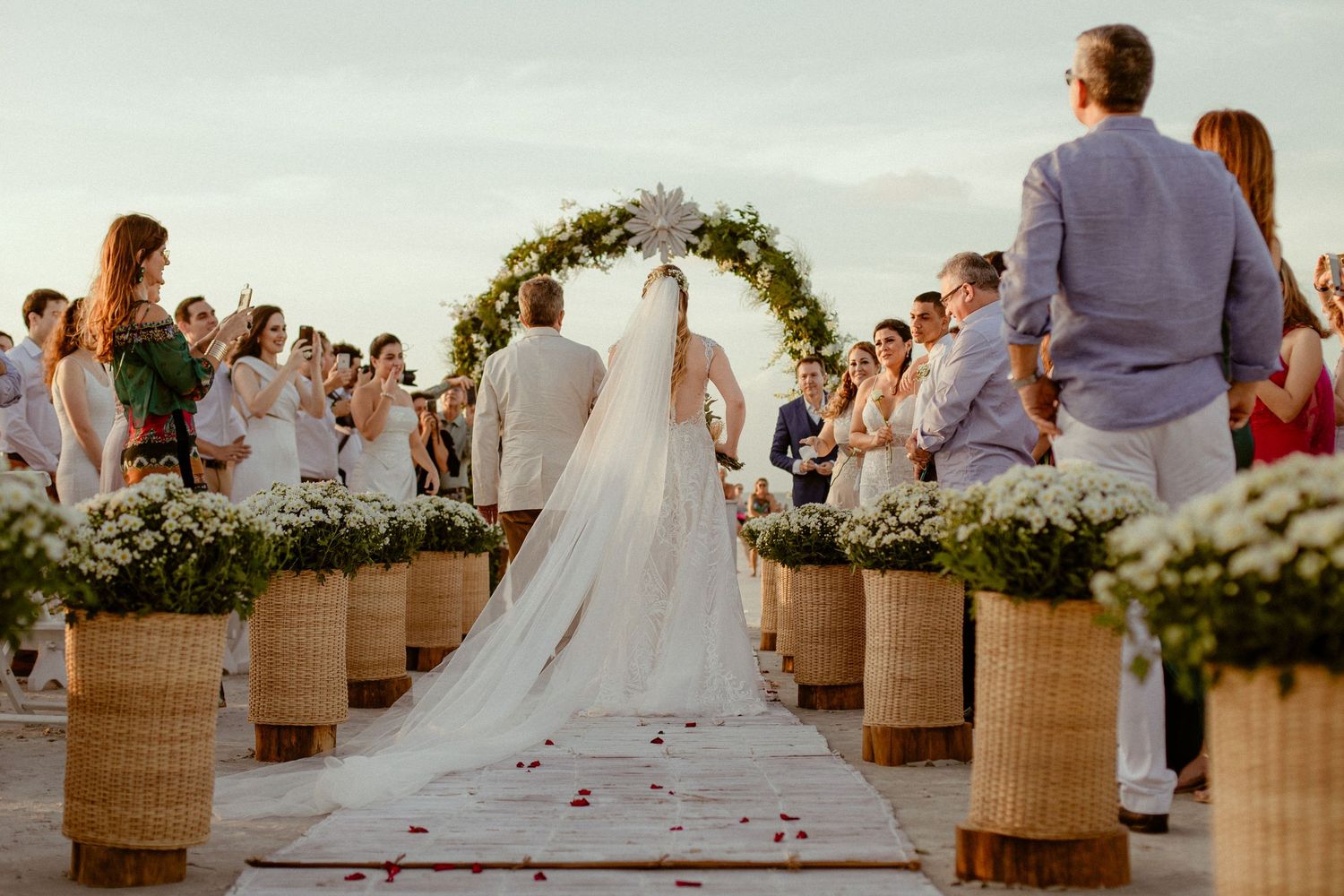 Outdoor beach wedding ceremony with floral arch and wicker basket aisle decorations during sunset.