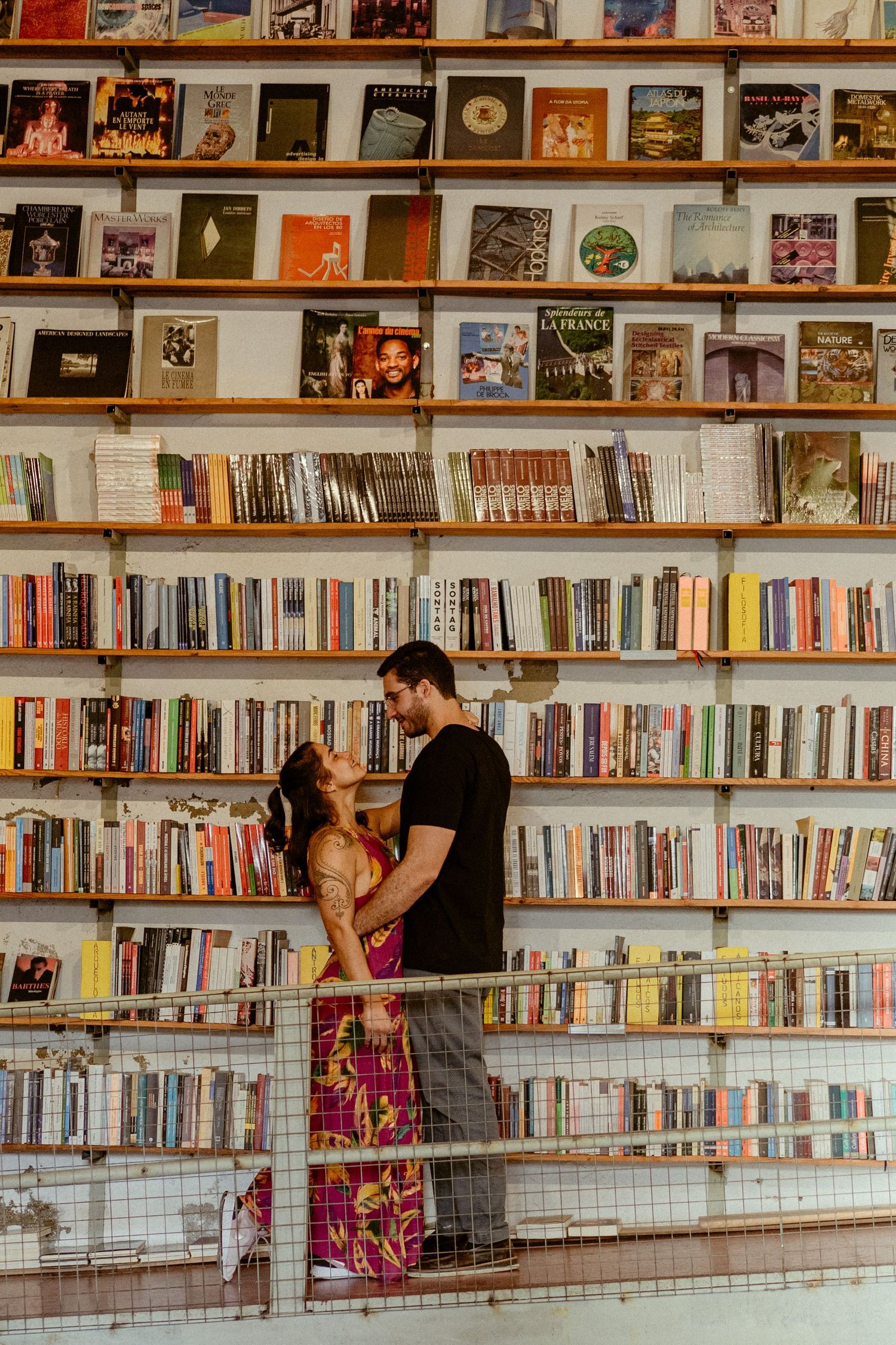A couple embraces against a wall of bookshelves filled with colorful books and albums in a library or bookstore.