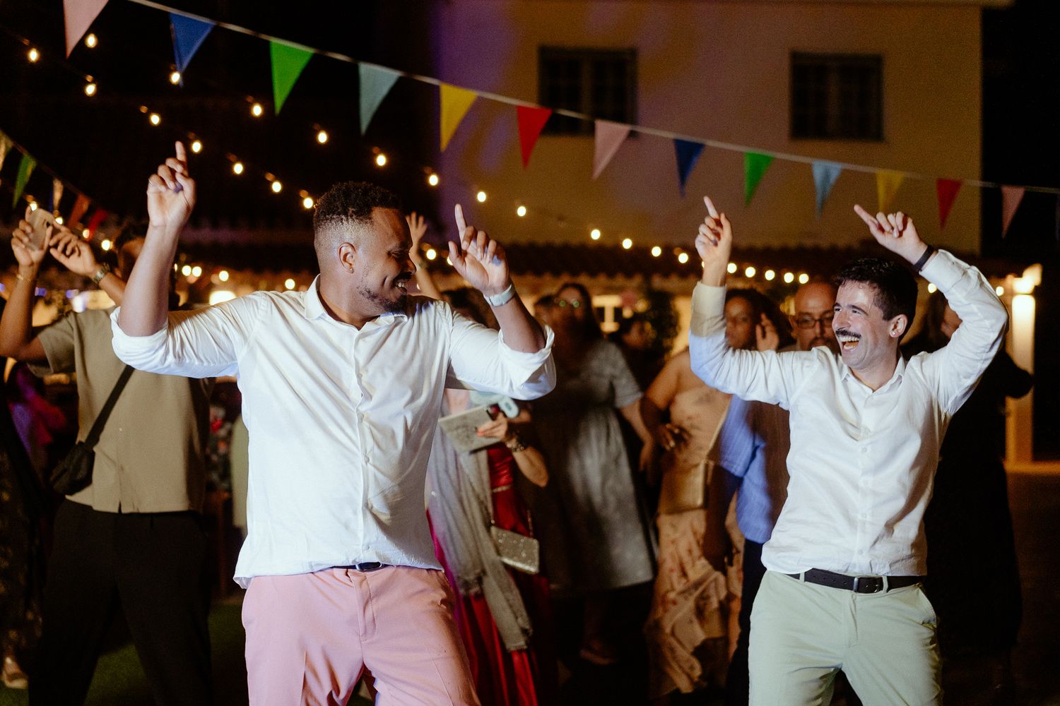 Two men dancing joyfully at an outdoor evening party with colorful bunting flags and string lights.