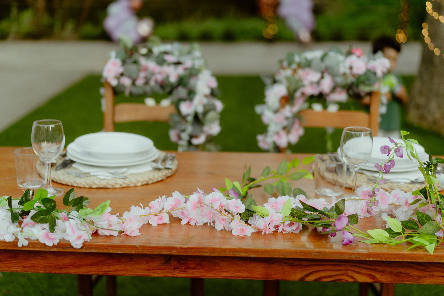 Elegant outdoor wedding table set with white plates, pink floral garlands, and glassware on wood table.