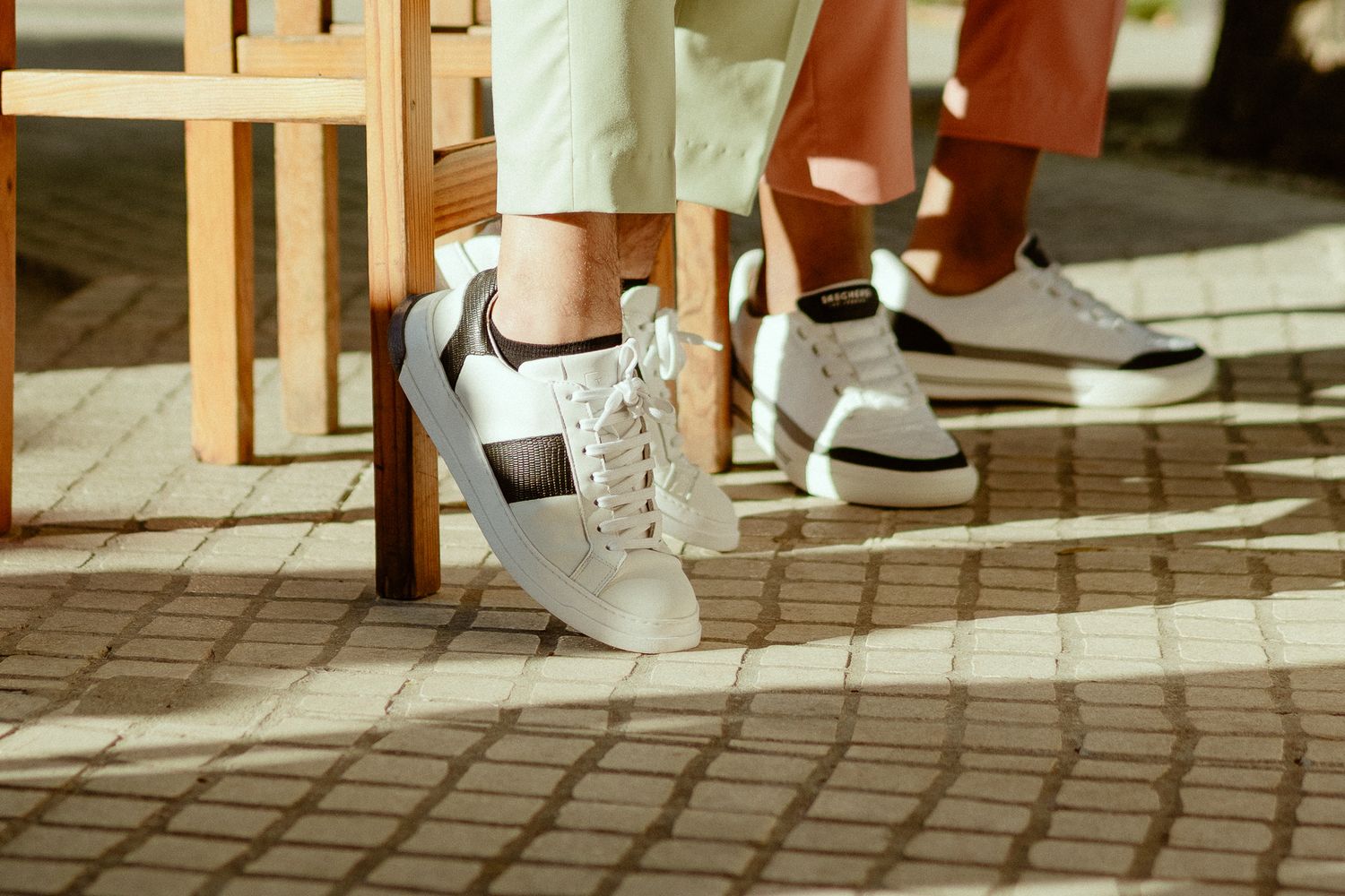 Two people wearing white leather sneakers with black accents sit outdoors on a sunny patio with brick flooring.