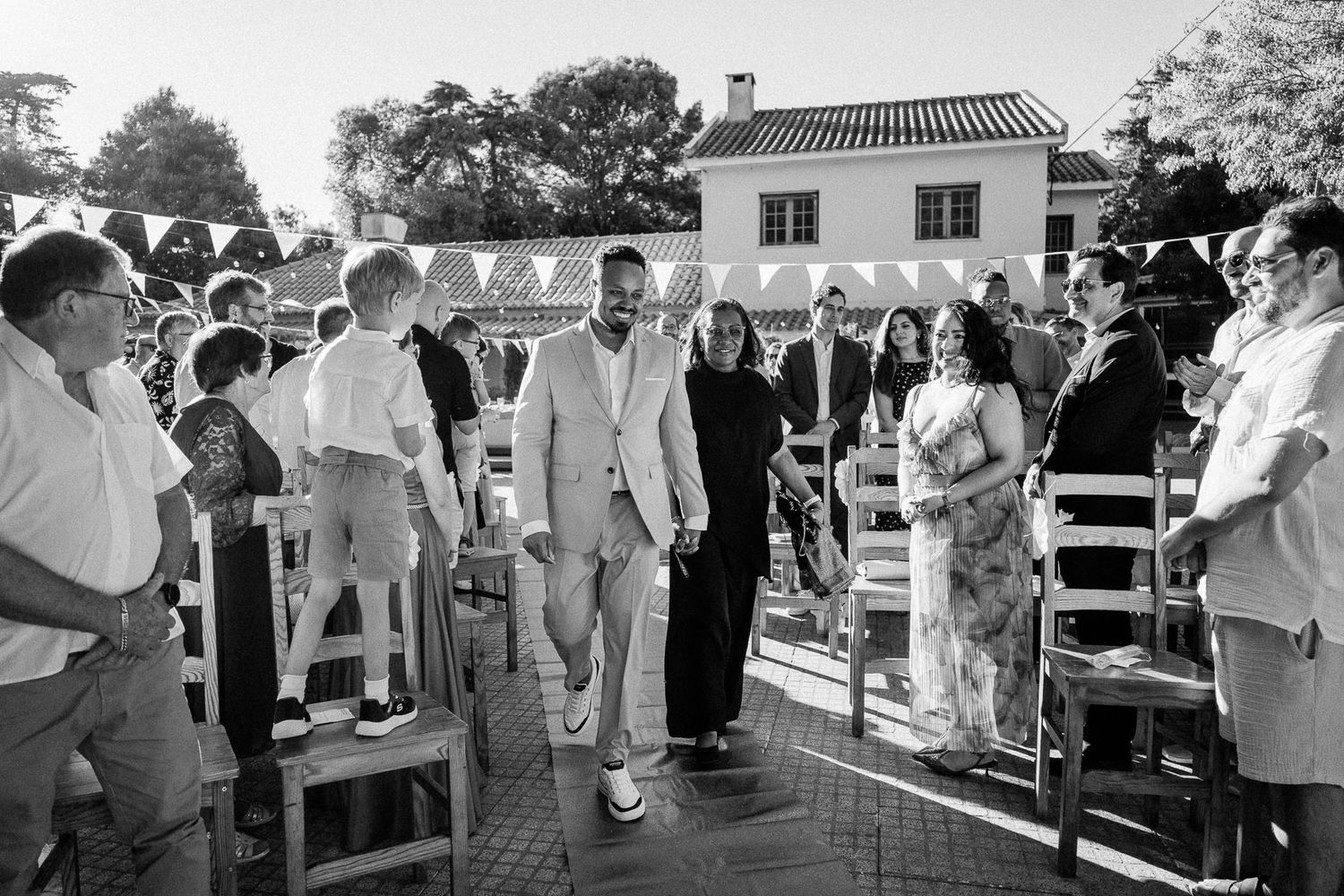 Black and white photo of a groom walking down the aisle at an outdoor wedding ceremony with guests seated.