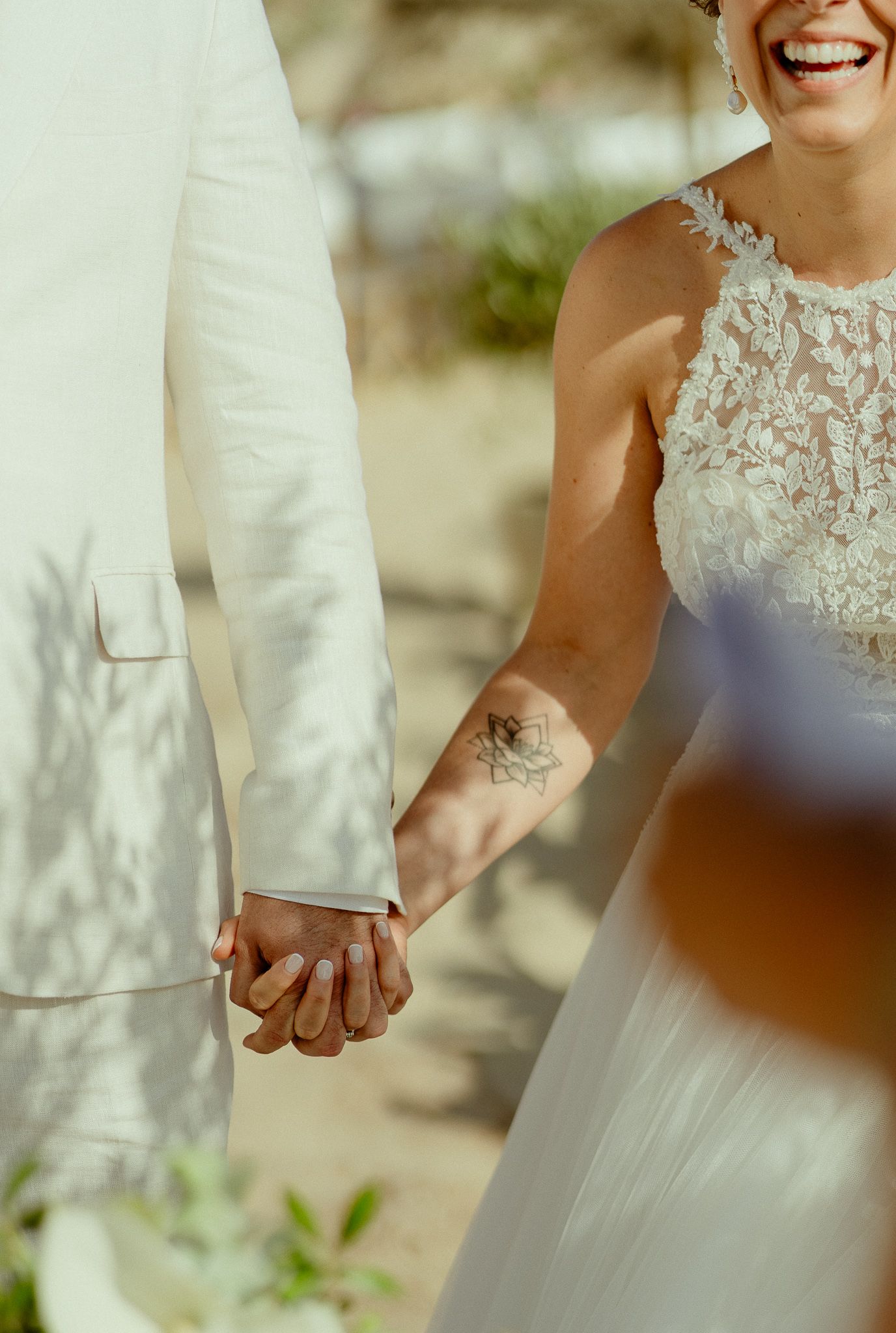 A laughing bride in lace gown holds hands with groom in white suit, her floral wrist tattoo visible.