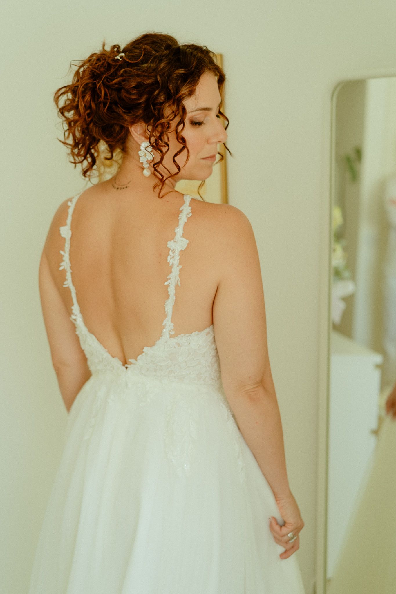 Bride in a backless white lace wedding gown with curly updo, standing near a mirror.