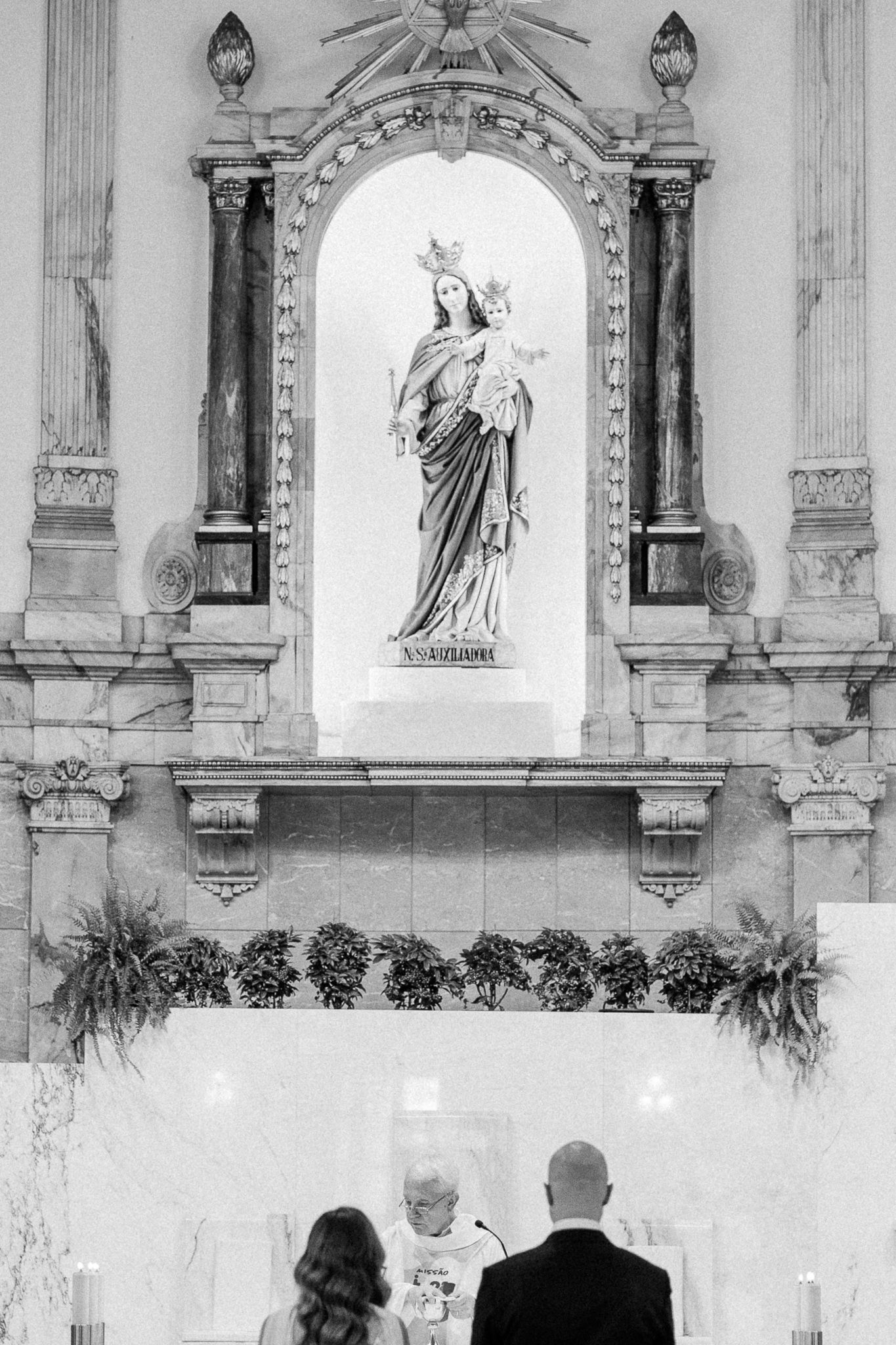 Black and white photo of a couple at a church altar with a statue of the Virgin Mary in the background.