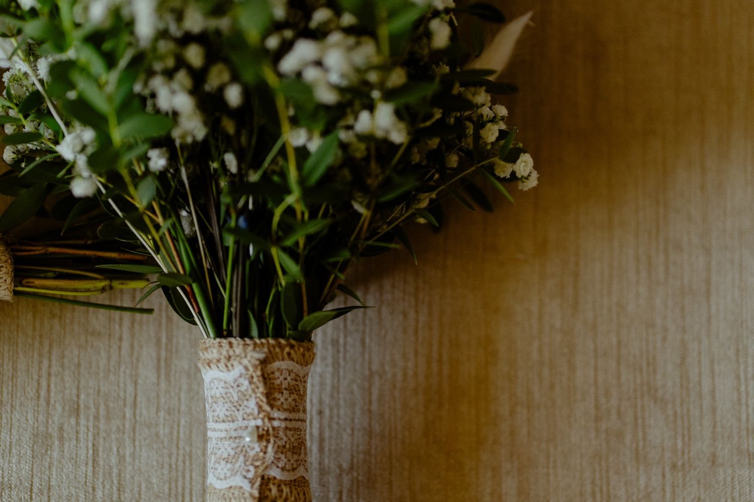 Elegant wedding bouquet with white flowers and greenery wrapped in lace ribbon against a neutral wall.