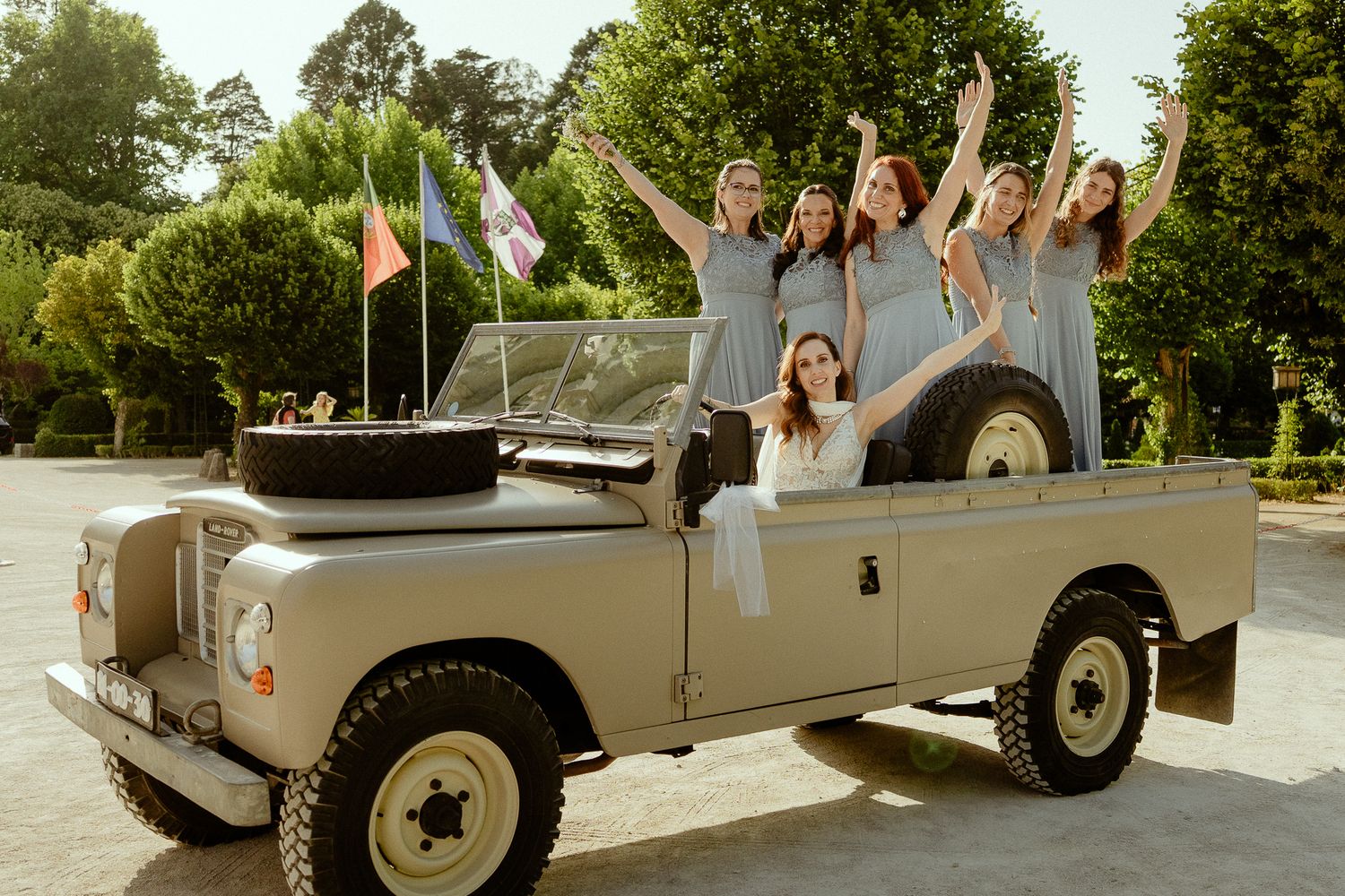 Bride and bridesmaids in gray dresses celebrating on a vintage Land Rover SUV at an outdoor wedding venue.