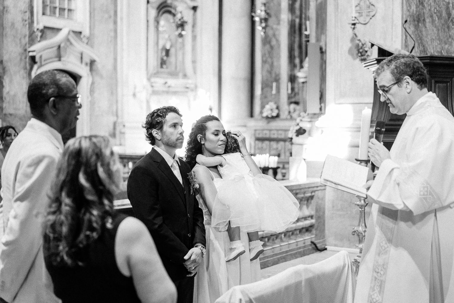 Black and white photo of a couple exchanging vows at a church altar with a priest during a formal wedding ceremony.