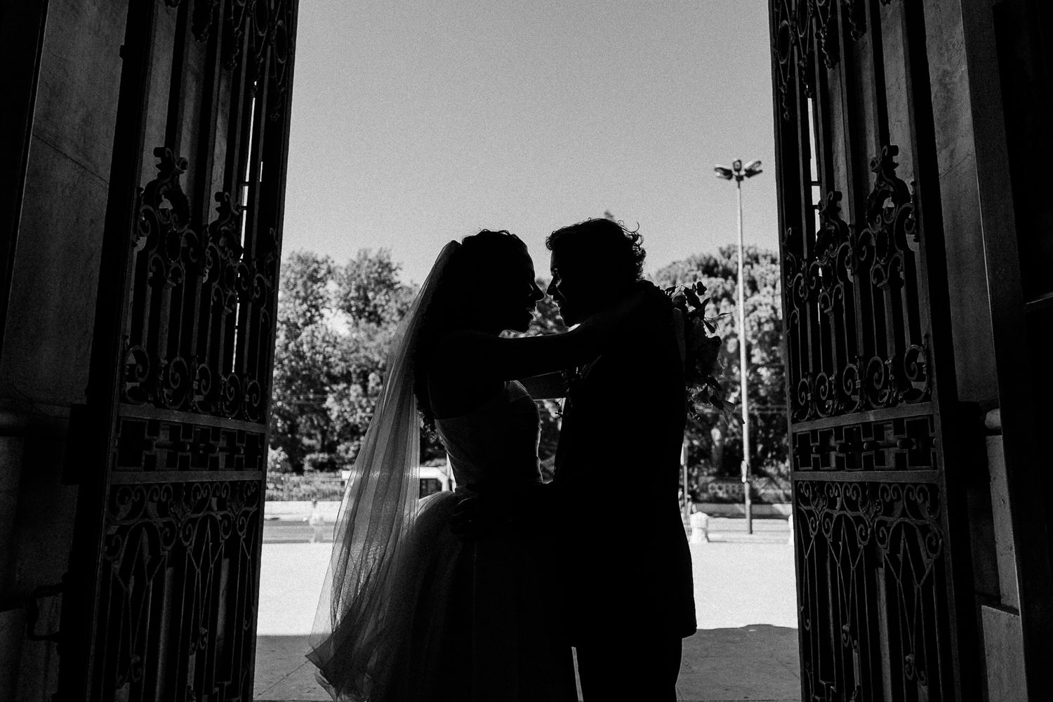 Black and white silhouette of bride and groom sharing romantic moment framed by ornate iron gates.