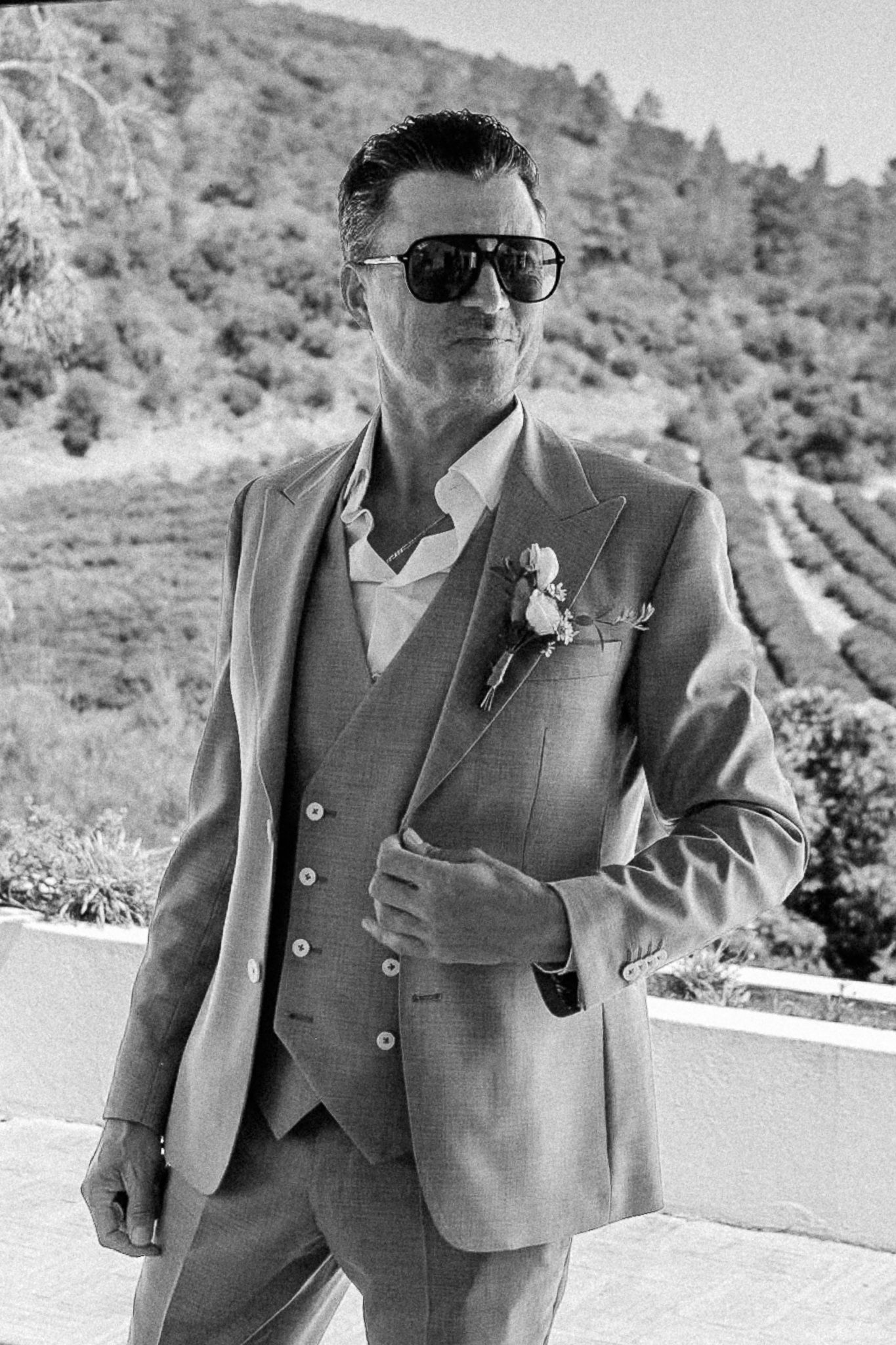 Stylish groom in a light gray three-piece suit with boutonniere and sunglasses outdoors.