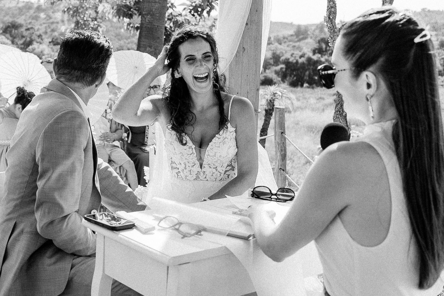 Black and white photo of a laughing bride in lace dress sharing a joyful moment with guests at an outdoor wedding.