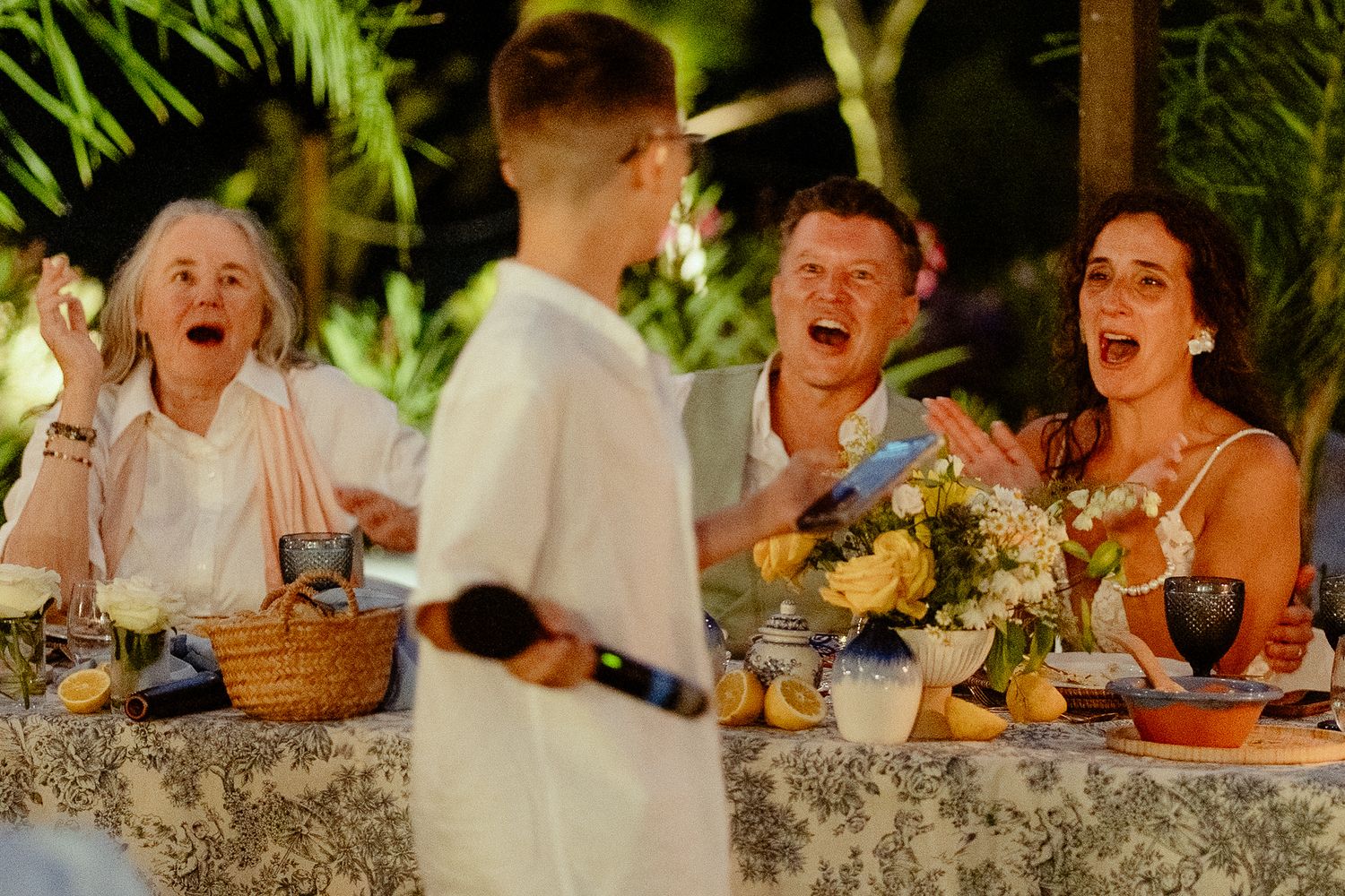Guests laugh joyfully at an outdoor dinner party with floral centerpieces and lush greenery.