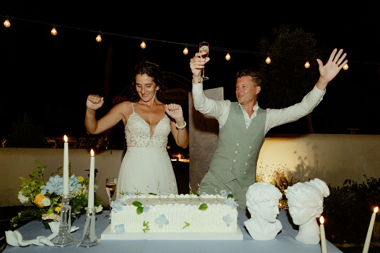 Bride and groom celebrate joyfully cutting their wedding cake outdoors at night, surrounded by candles and floral arrangements.