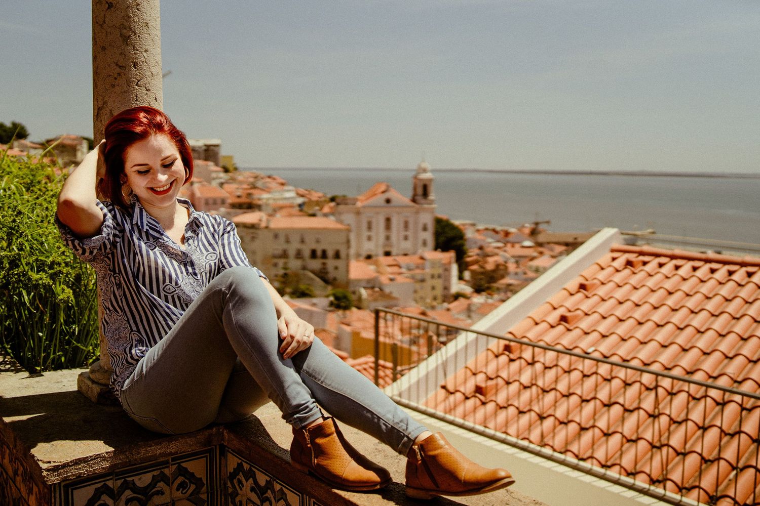 Woman relaxing on rooftop overlooking coastal town with terracotta tiles and ocean view.