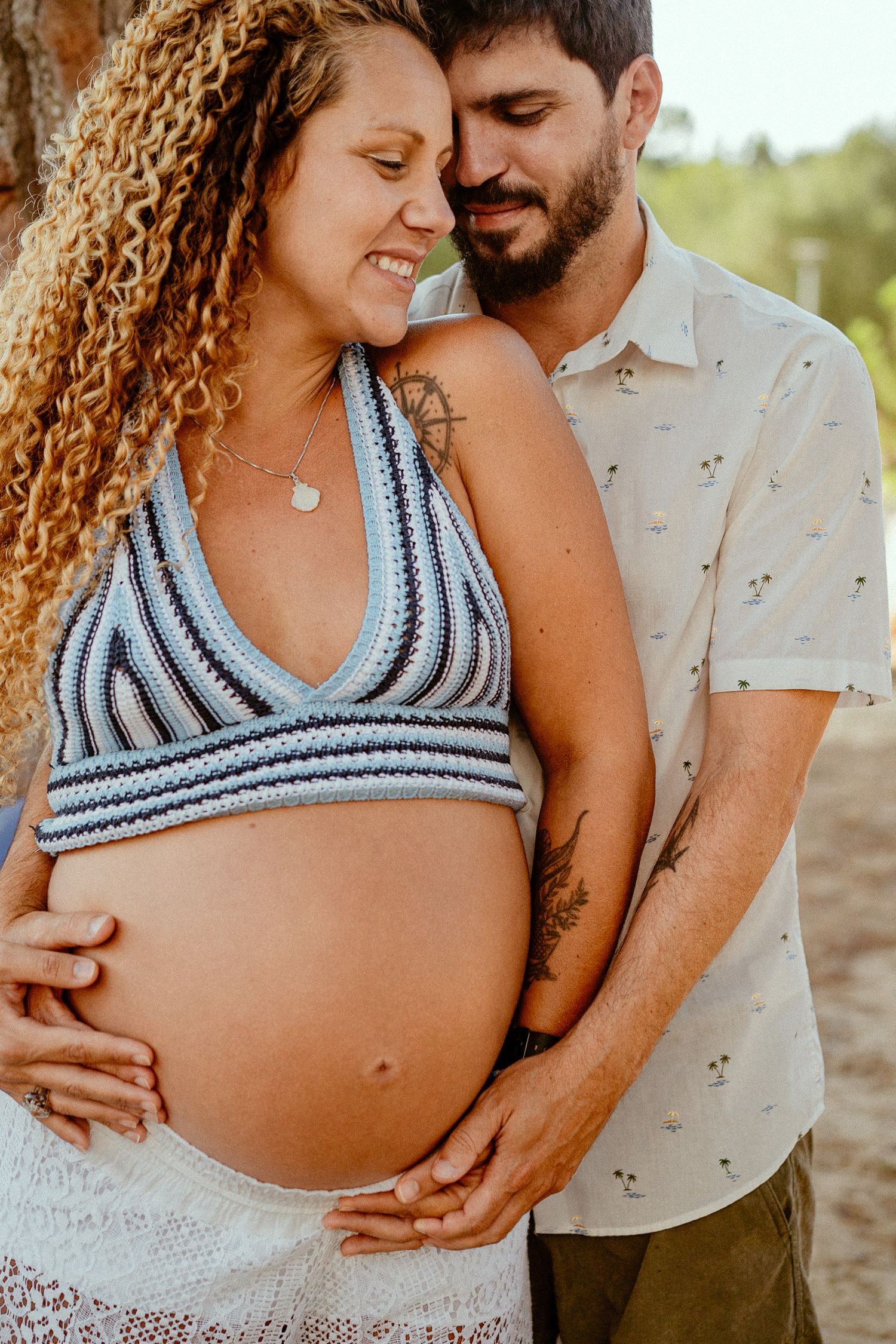 A tender maternity photo showing an expectant couple embracing outdoors, the mother wearing a blue striped crop top.