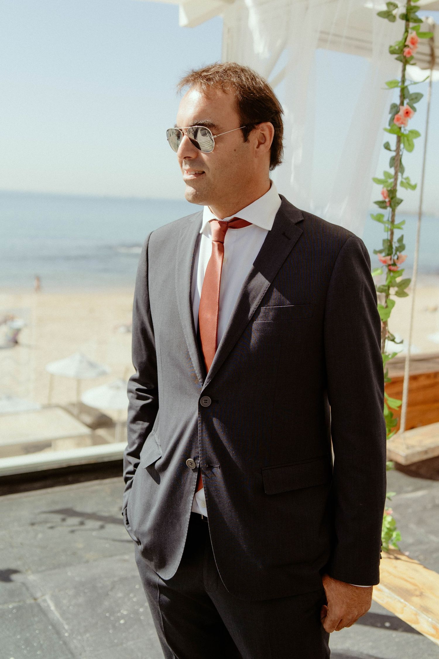 A person in a dark suit and coral tie stands outdoors wearing sunglasses with an ocean view in the background.