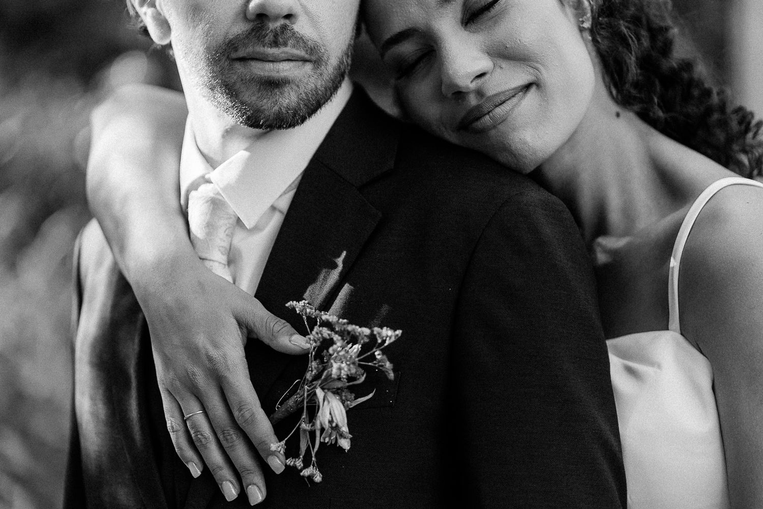 Black and white wedding photo of bride resting head on groom's shoulder, showcasing boutonniere and elegant attire.
