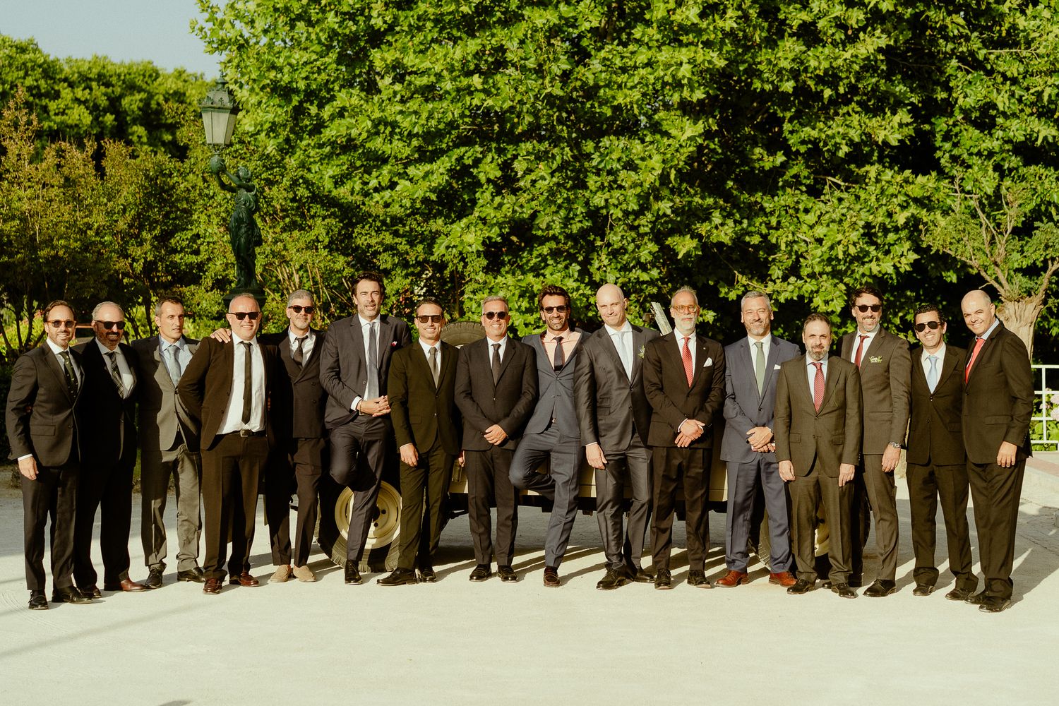 Large group of well-dressed men in suits posing together outdoors near green trees on a sunny day.
