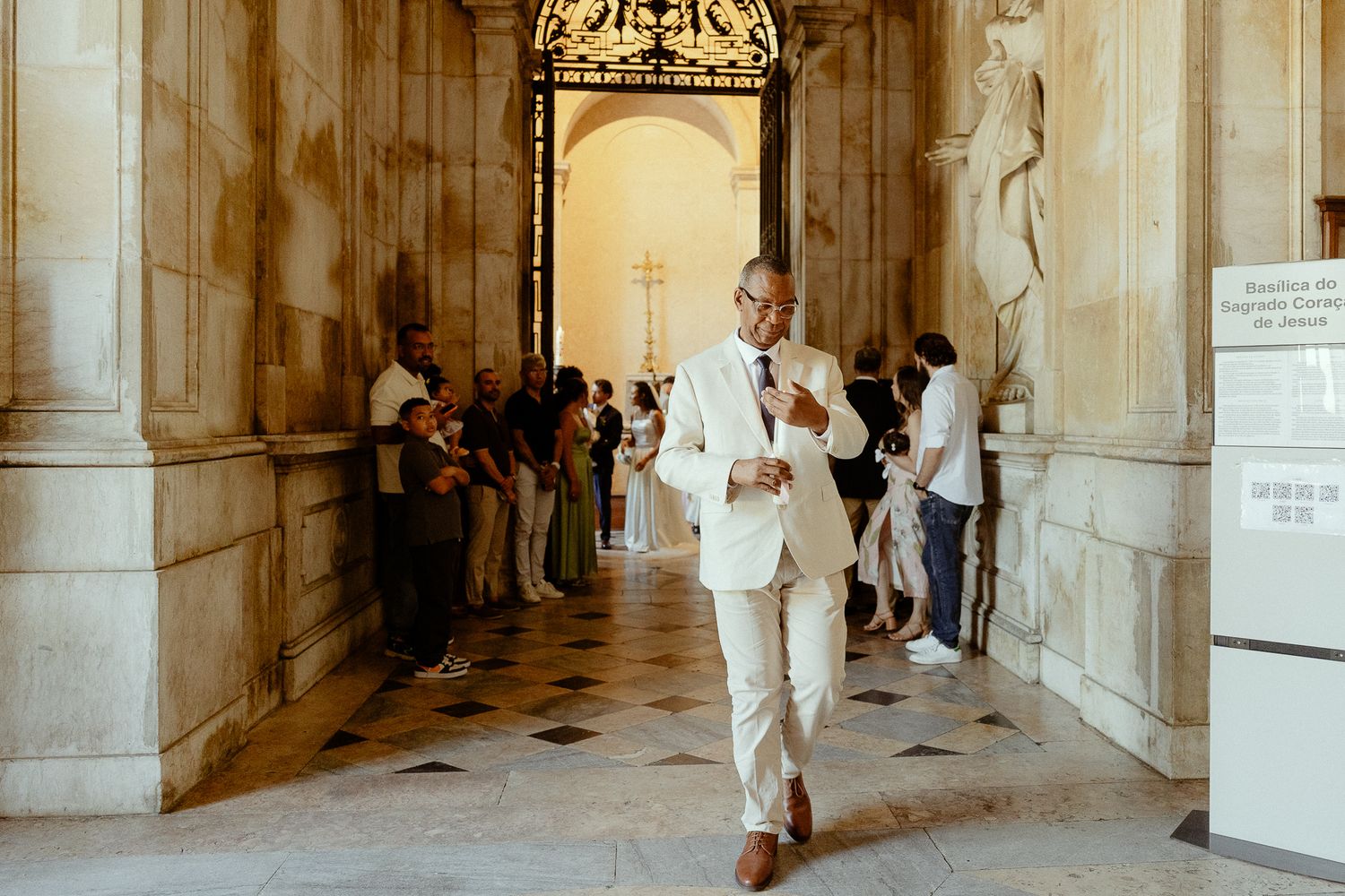 Elegant man in white suit walking through ornate marble hall with visitors in background.