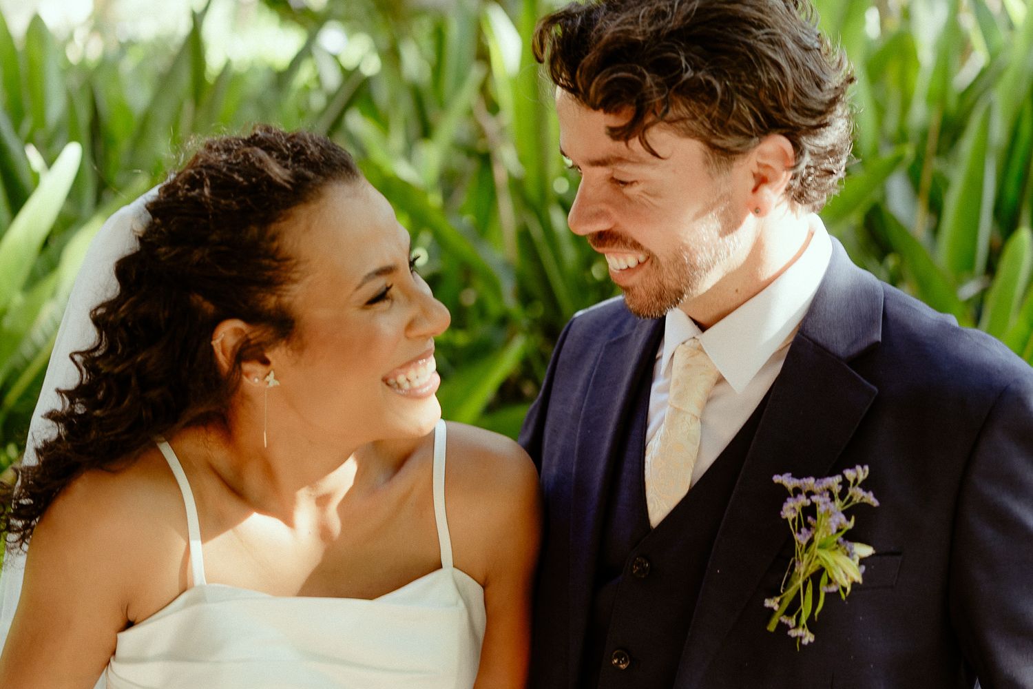 Happy bride and groom smiling at each other on their wedding day, groom wearing navy suit with floral boutonniere.