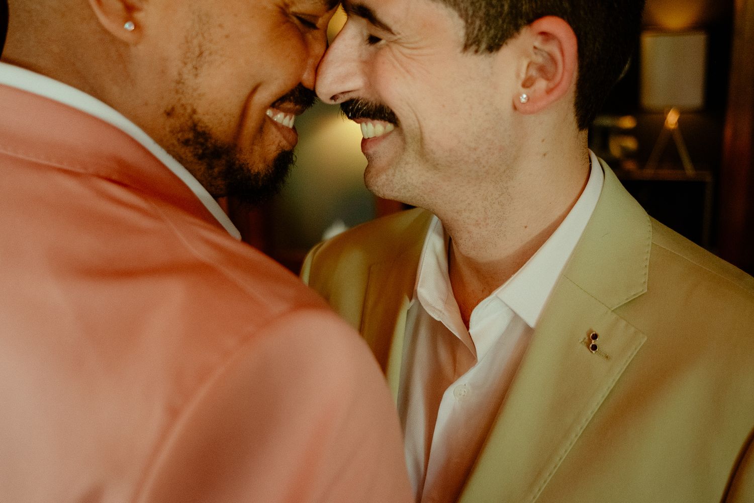 Two men in colorful suits share an intimate moment, smiling and leaning foreheads together at a celebration.