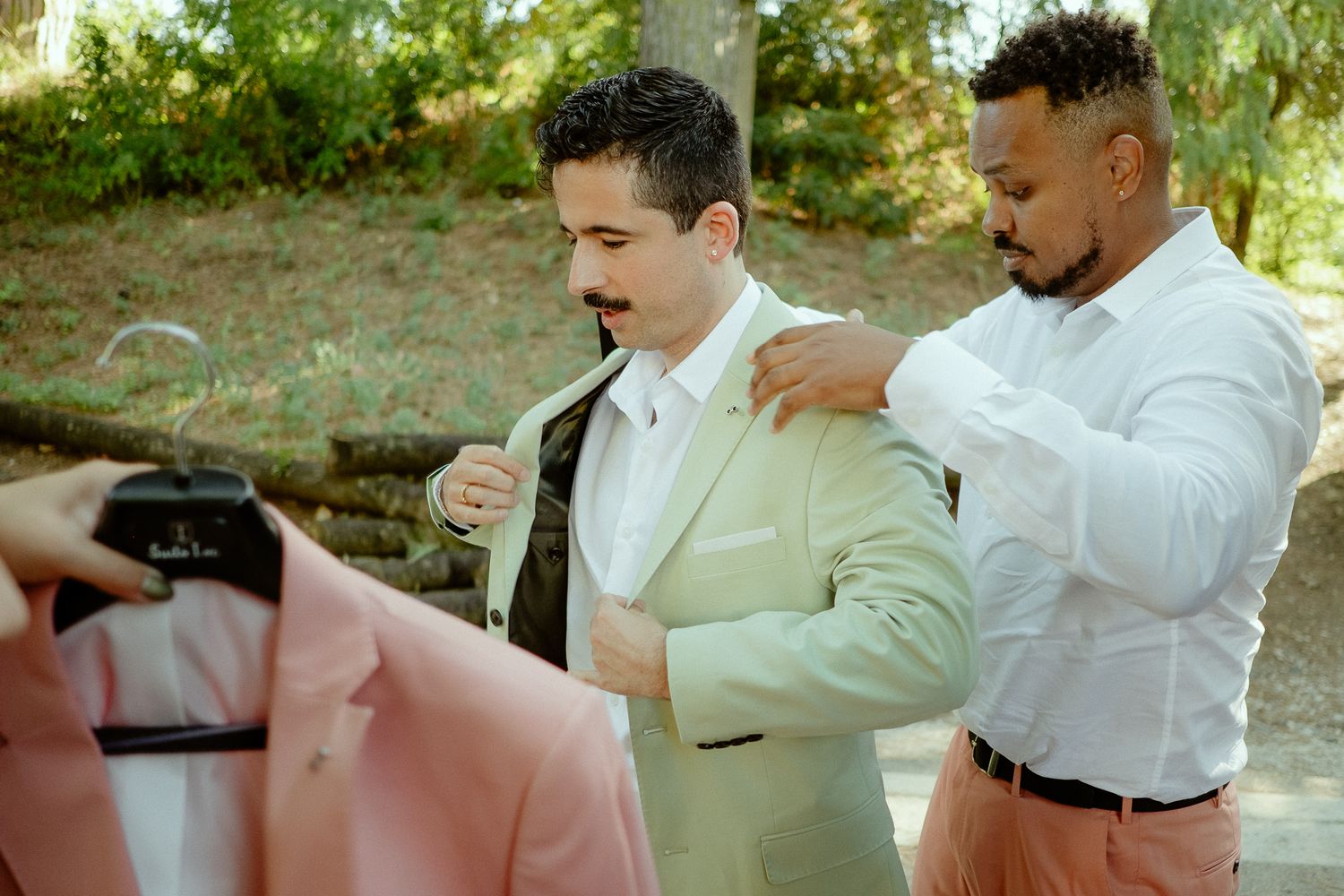 Groom in sage green suit being helped by groomsman outdoors, with pink suit jacket visible in foreground.