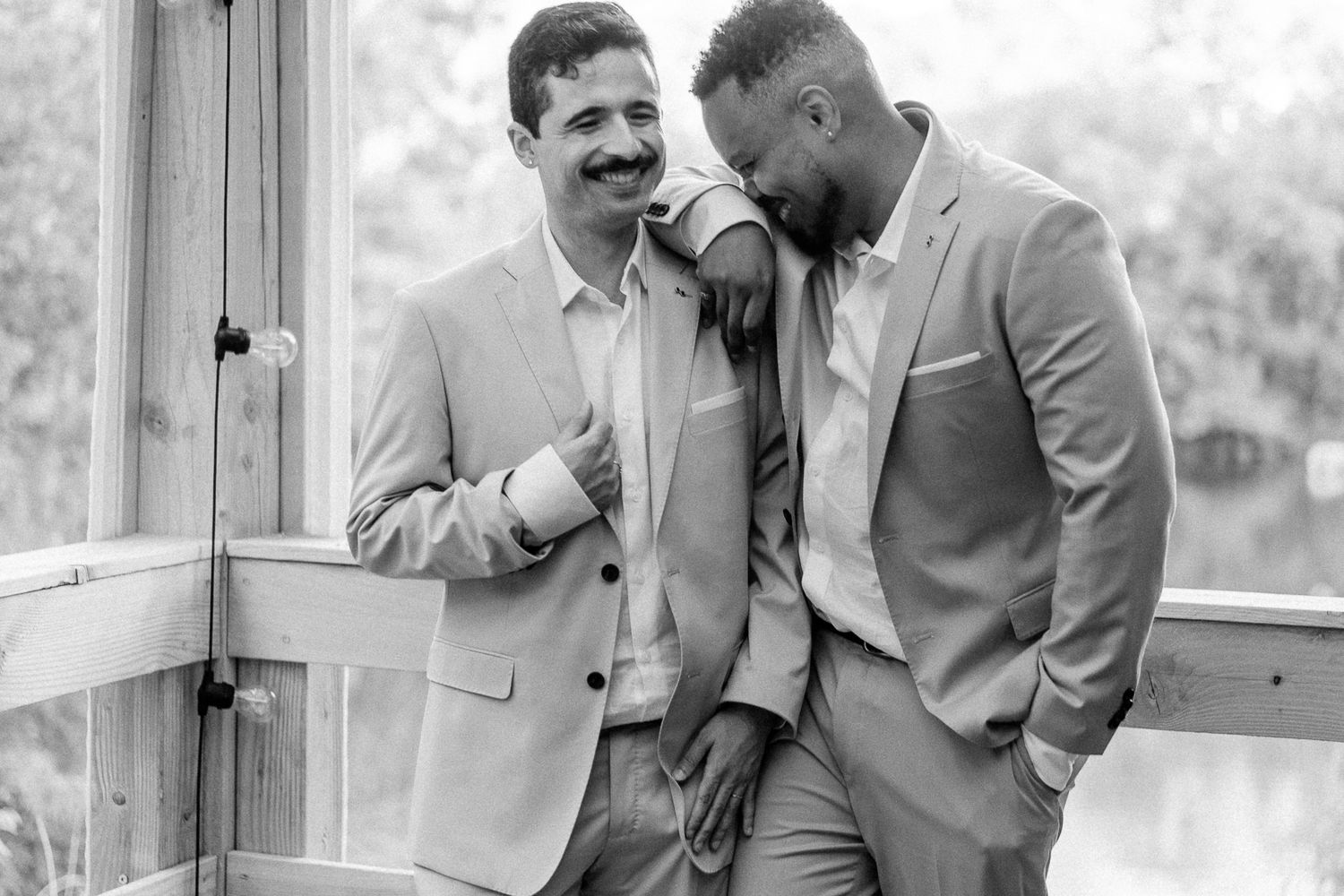 Two men in matching light suits share a joyful moment together, laughing and leaning on each other in a black and white photo.