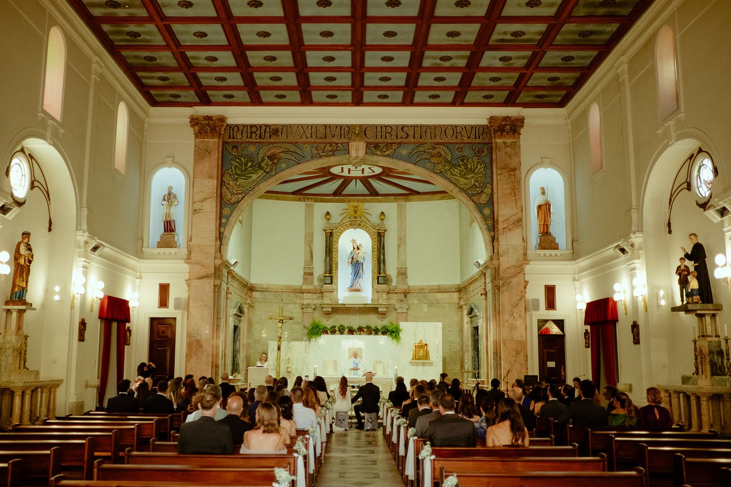 Elegant Catholic church wedding ceremony with ornate altar, wooden pews, and guests gathered under decorative arched ceiling.