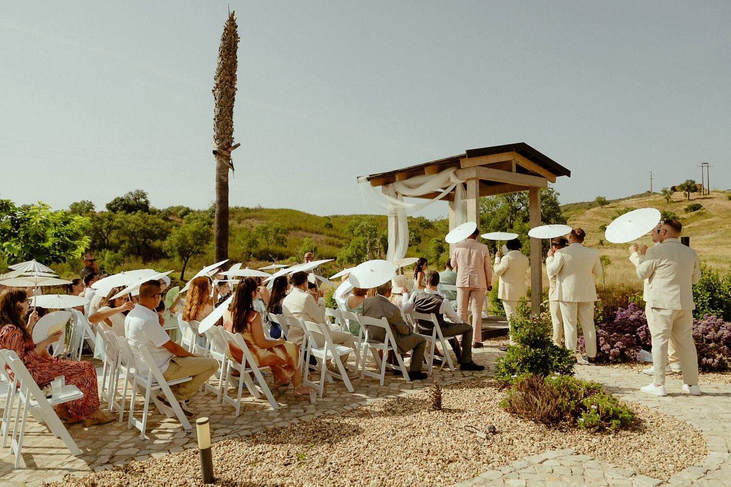 Outdoor wedding ceremony with guests in white attire seated on white chairs under sunny skies at a coastal venue.