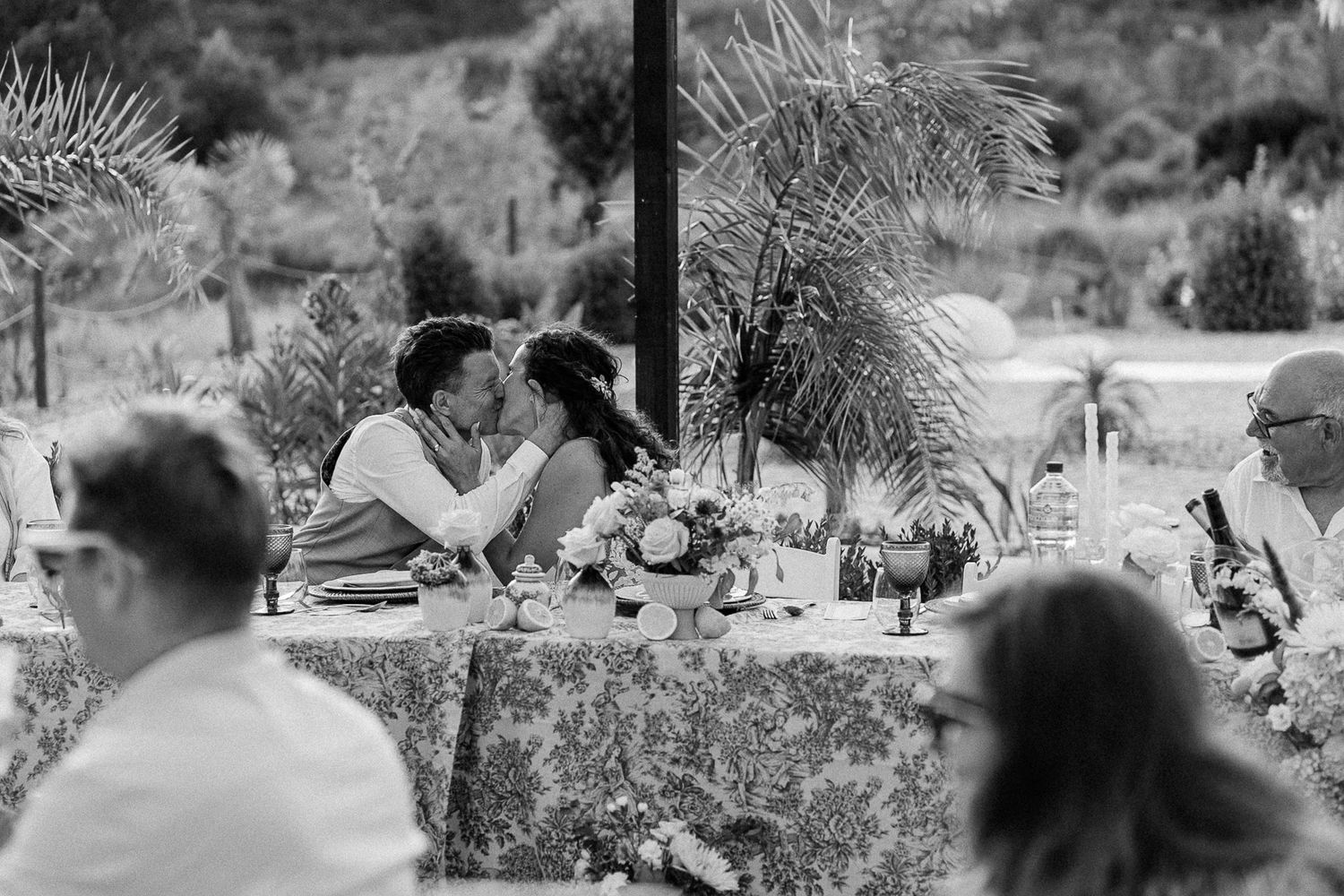 Black and white wedding reception photo of couple sharing a kiss at the head table with guests seated nearby.