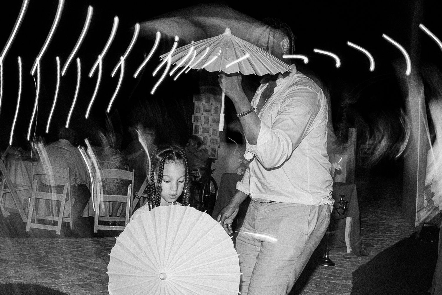 Black and white photo of a child and adult holding decorative parasols at an outdoor evening event.