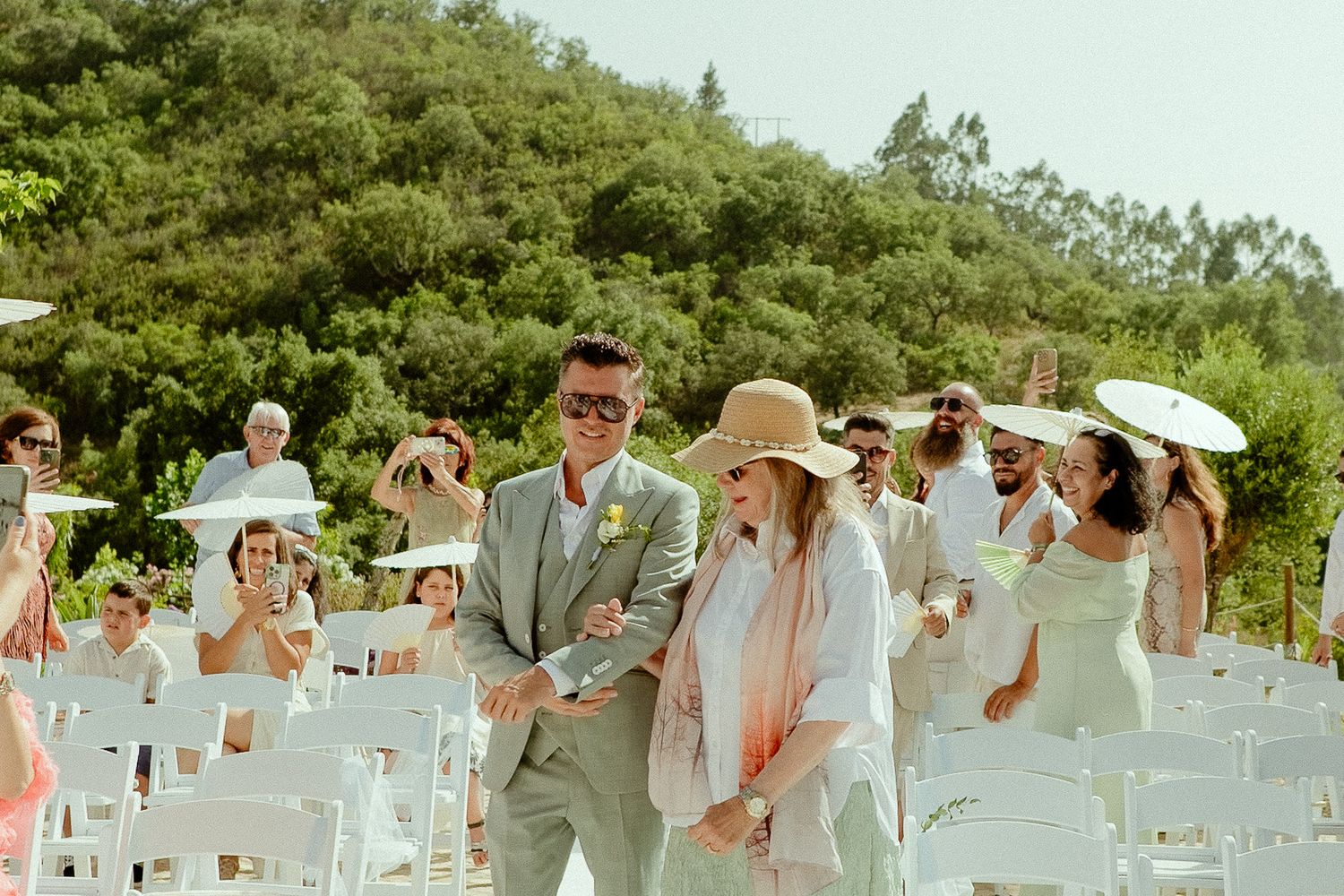 Groom in tan suit walks down outdoor wedding aisle with woman in pink shawl and sun hat, white chairs and greenery.
