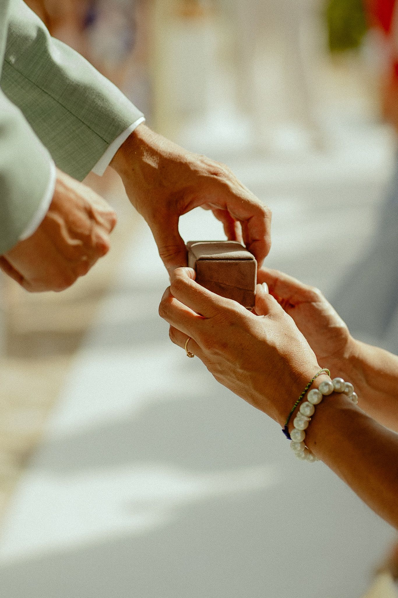 Close-up of hands exchanging a ring box during a wedding ceremony, with one person wearing a pearl bracelet.