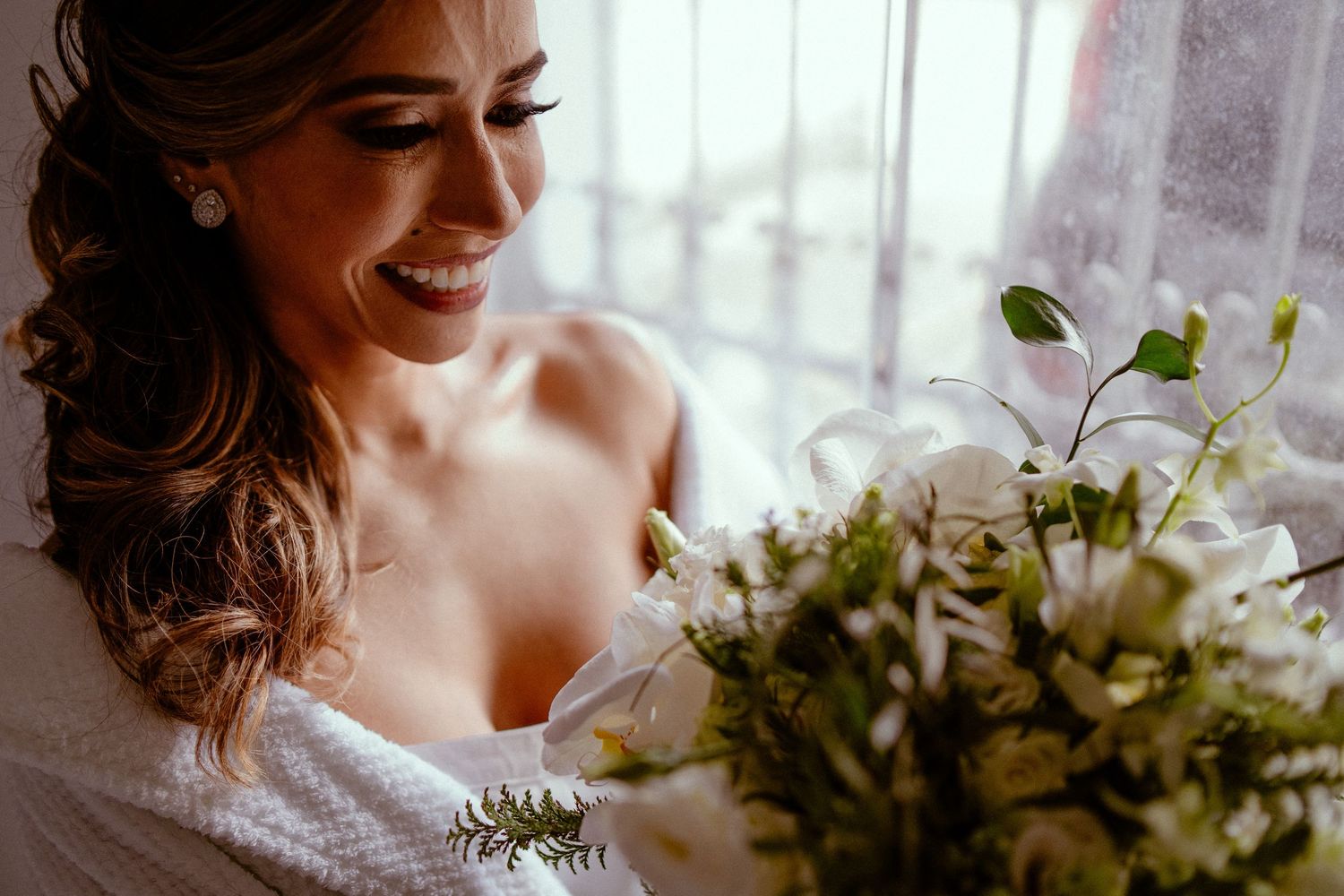 A bride in a white wedding dress smiles while holding a lush green and white floral bouquet by a window.