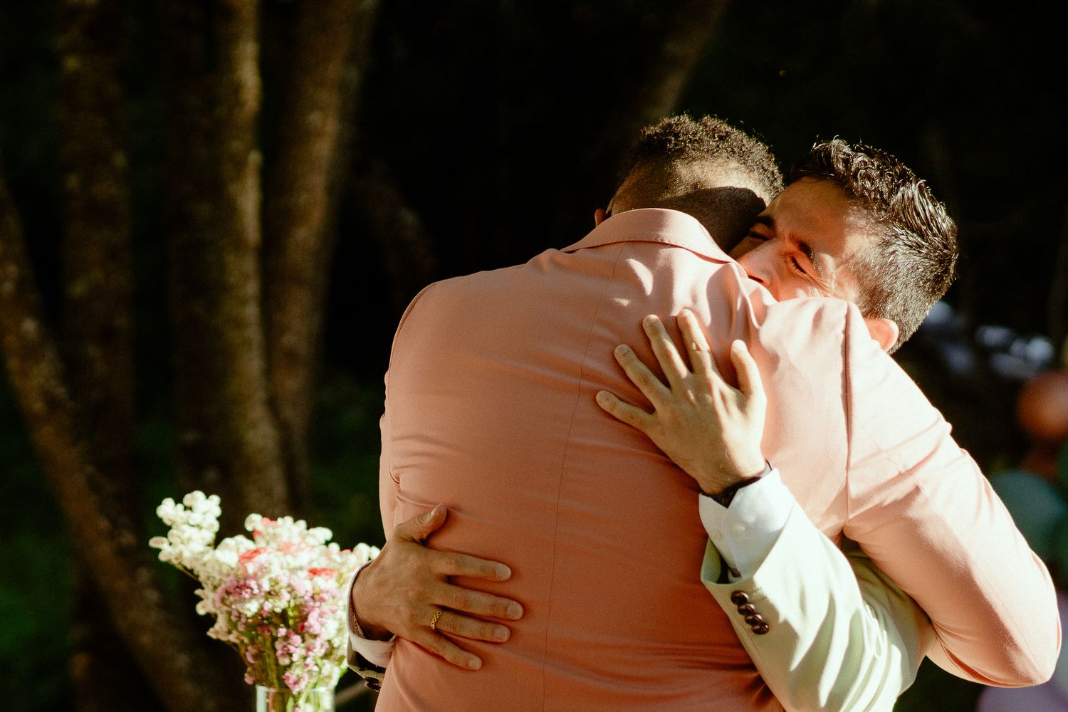 Two grooms share an emotional embrace during their outdoor wedding ceremony, one wearing a pink suit holding a floral bouquet.