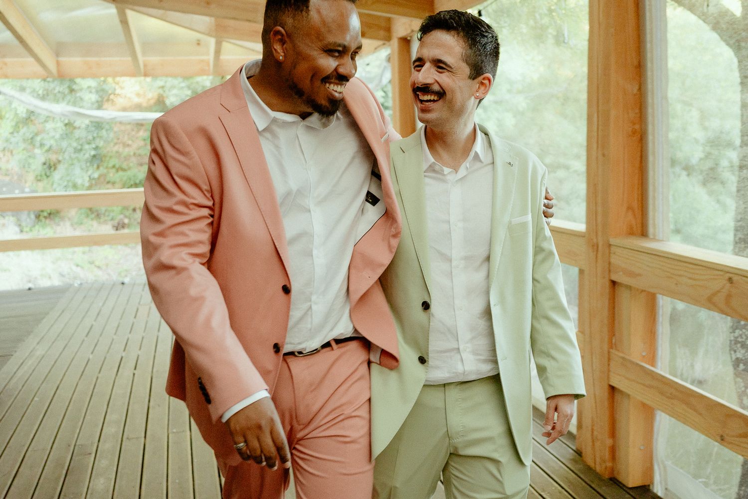 Two men in pastel suits laughing and walking together on a wooden deck, sharing a joyful moment.