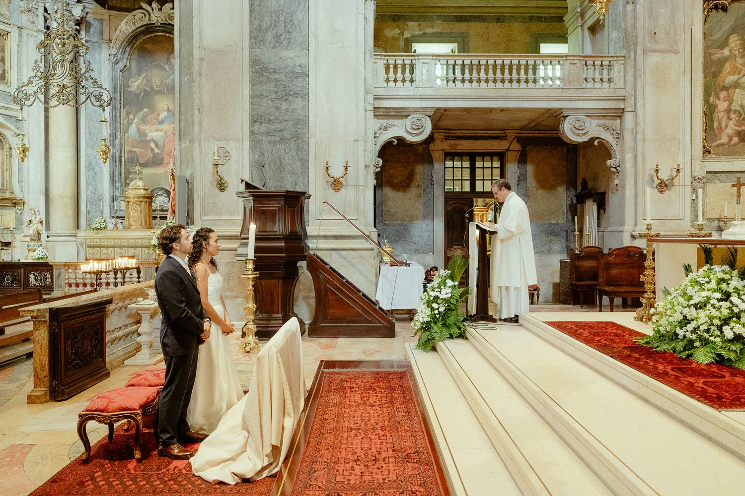 Elegant wedding ceremony in an ornate church with bride and groom standing before an officiant at the altar.