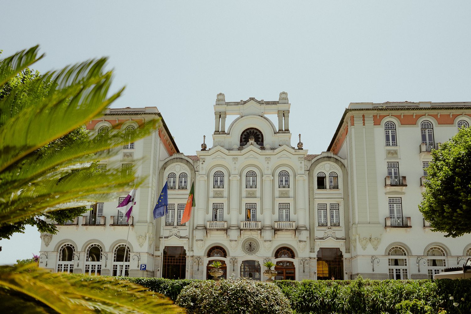 Grand white neoclassical building facade with flags and lush green trees in foreground.