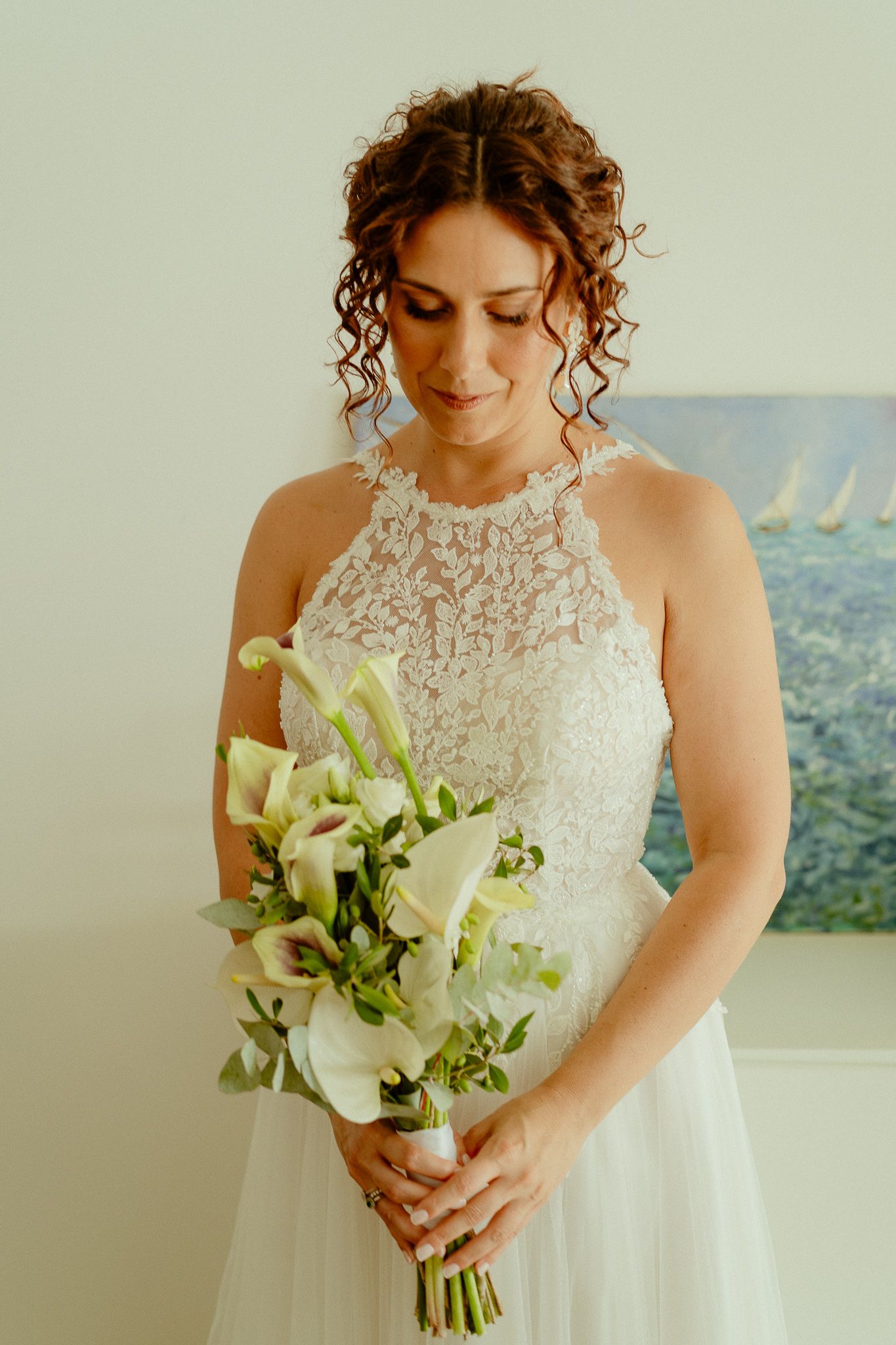Bride in lace halter wedding gown holding white calla lily bouquet, looking down with curly hair and soft smile.