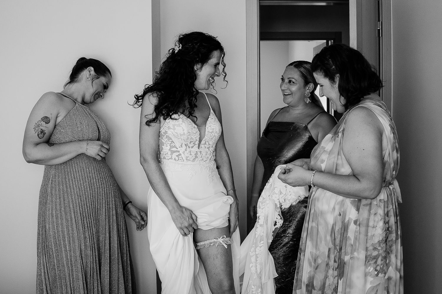 Black and white photo of bride in wedding dress with bridesmaids helping her prepare in a bright room.