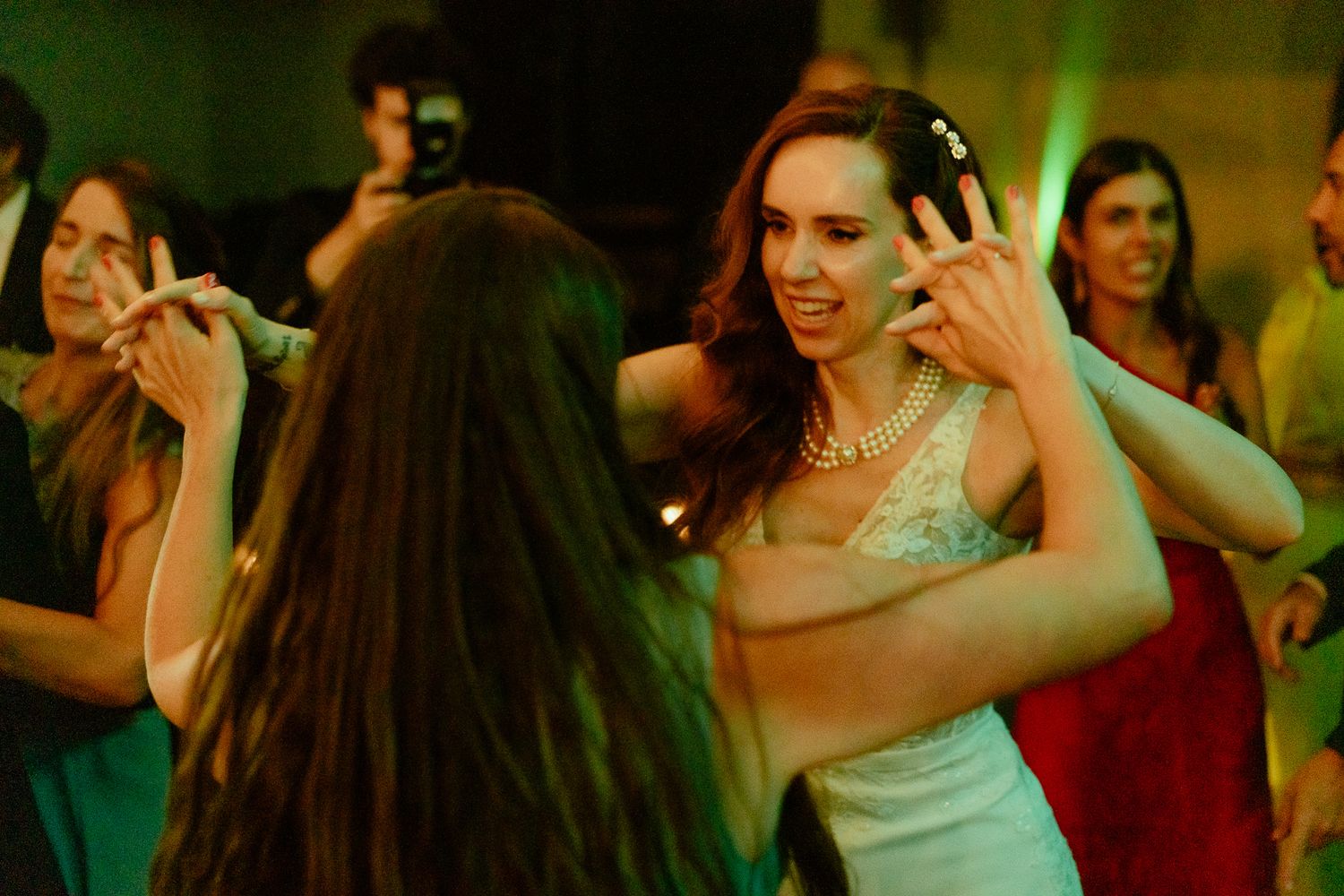 Women dancing at a party, one wearing a white dress and pearl necklace, making hand gestures under green lights.