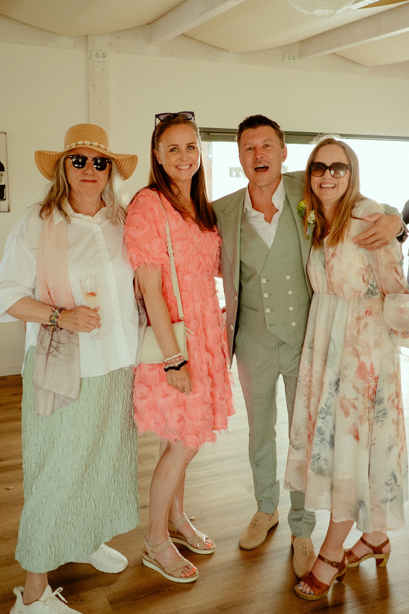 Four elegantly dressed guests smiling together at a sunny wedding celebration, wearing pastel outfits and summer accessories.