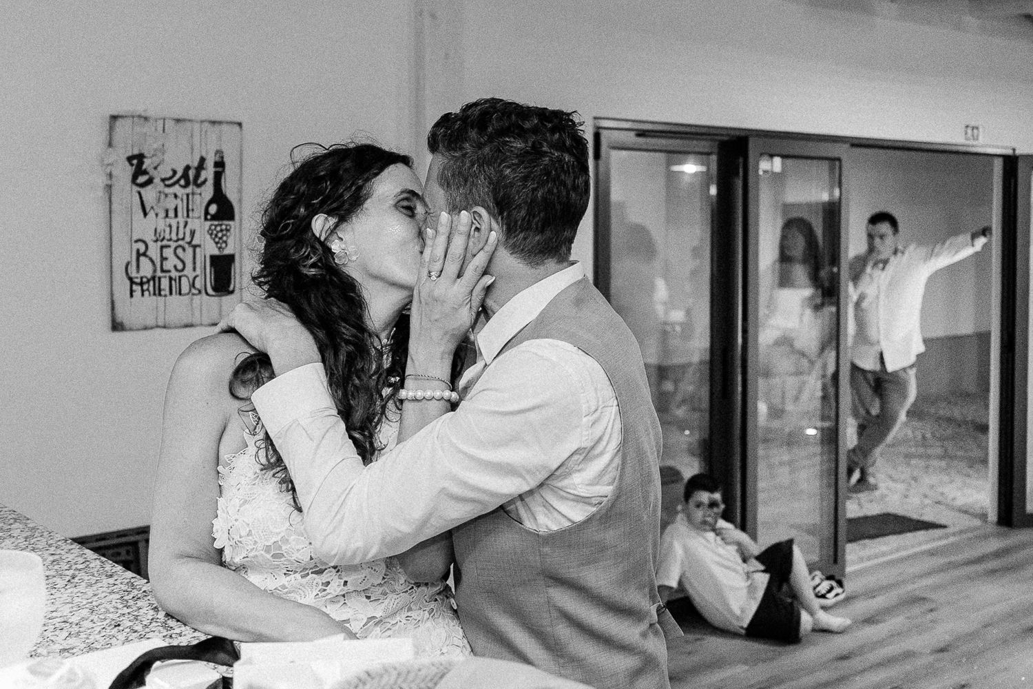 Black and white wedding photo of couple kissing while guests celebrate in background through glass door.